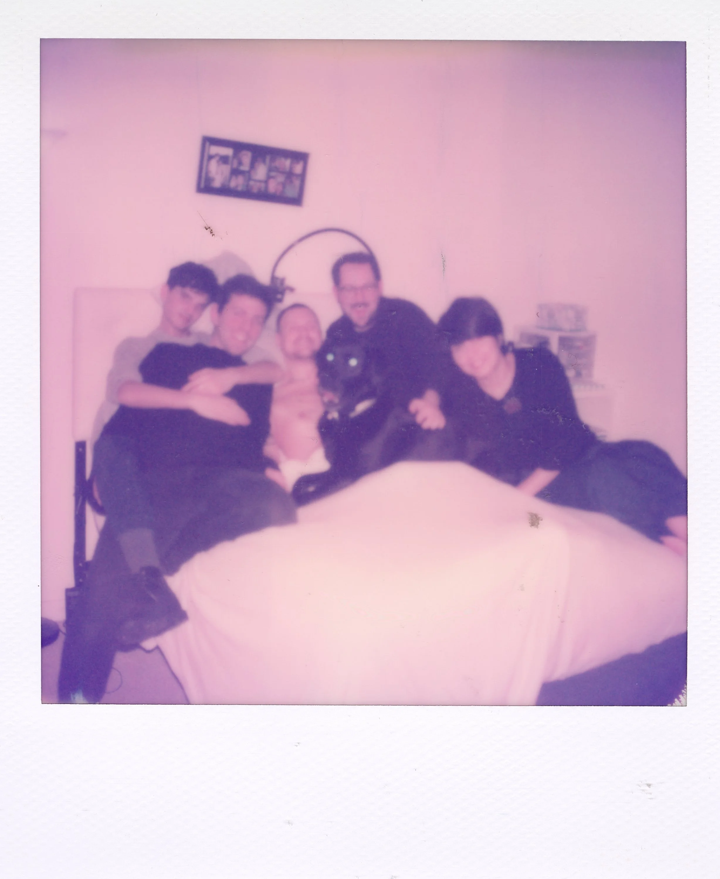 Group of five people sitting and lying on a bed, smiling and hugging, with a black dog in the center. The room has a pink wall, a framed picture above, and a nightstand with books on the right.