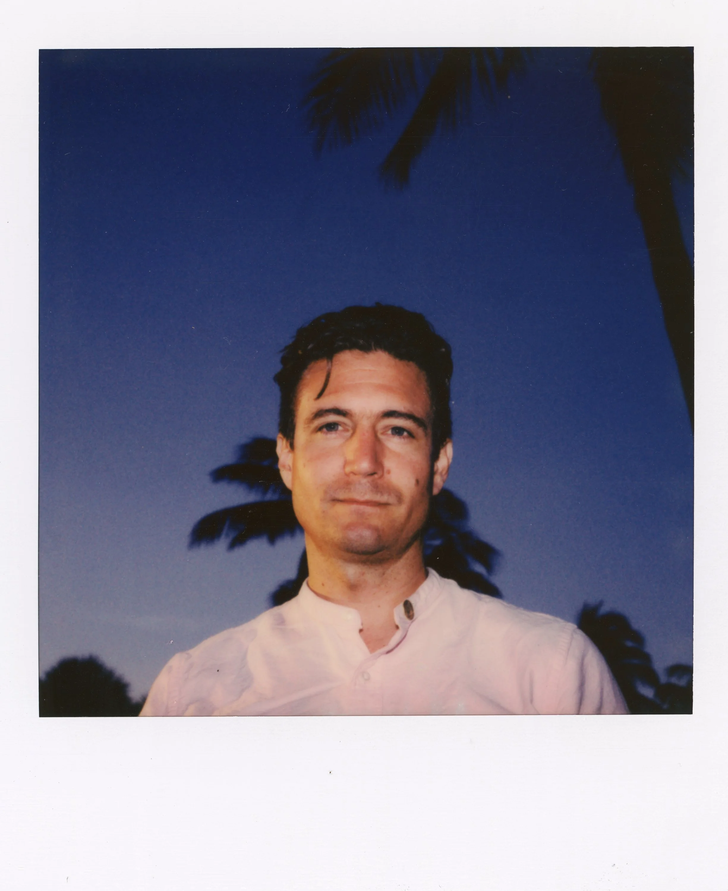 A man with dark hair and a light-colored shirt standing outdoors at dusk, with palm trees silhouette against a dark blue sky in the background.