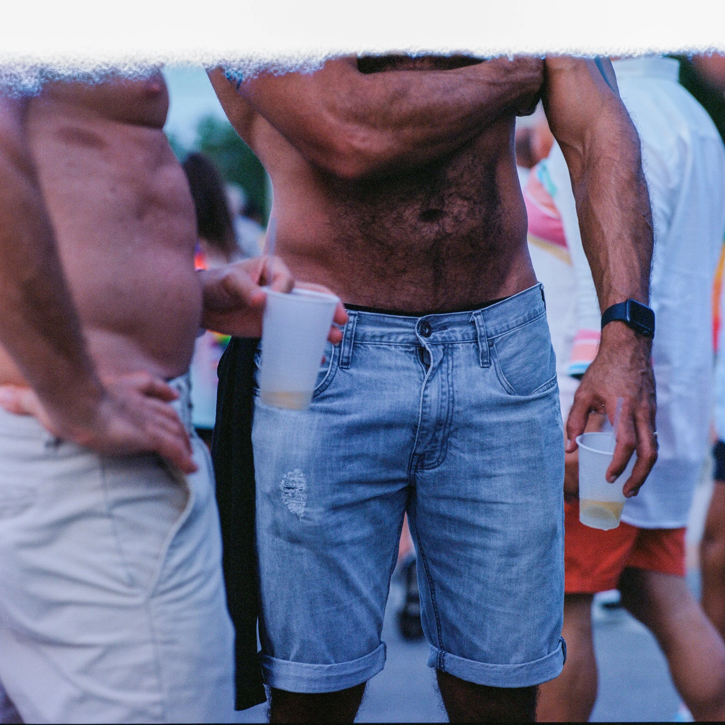 A group of people at an outdoor event, focusing on a shirtless man wearing denim shorts and a smartwatch, holding a plastic cup with a beverage.