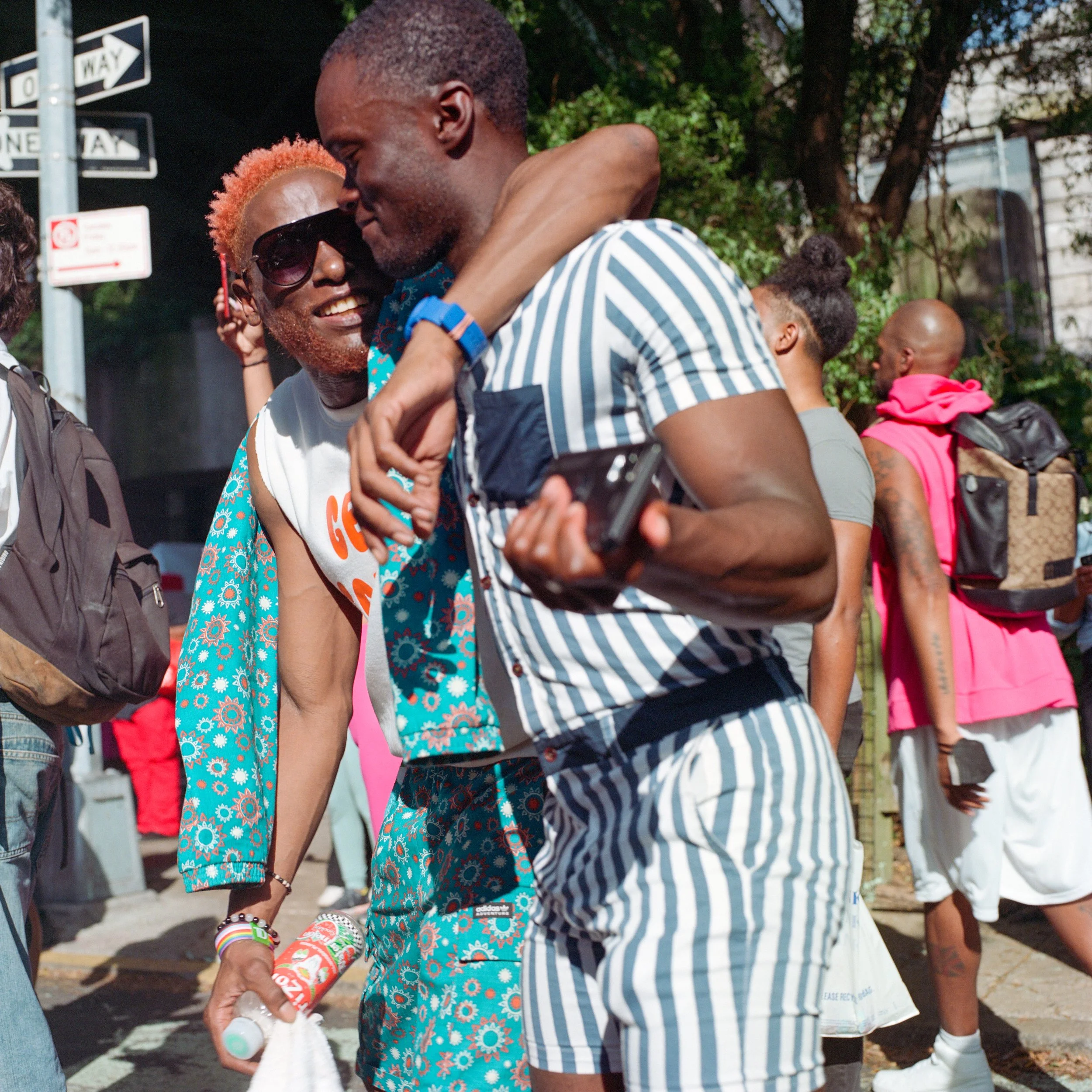 Two young men hugging and smiling at each other outdoors on a sunny day, with several other people in the background.