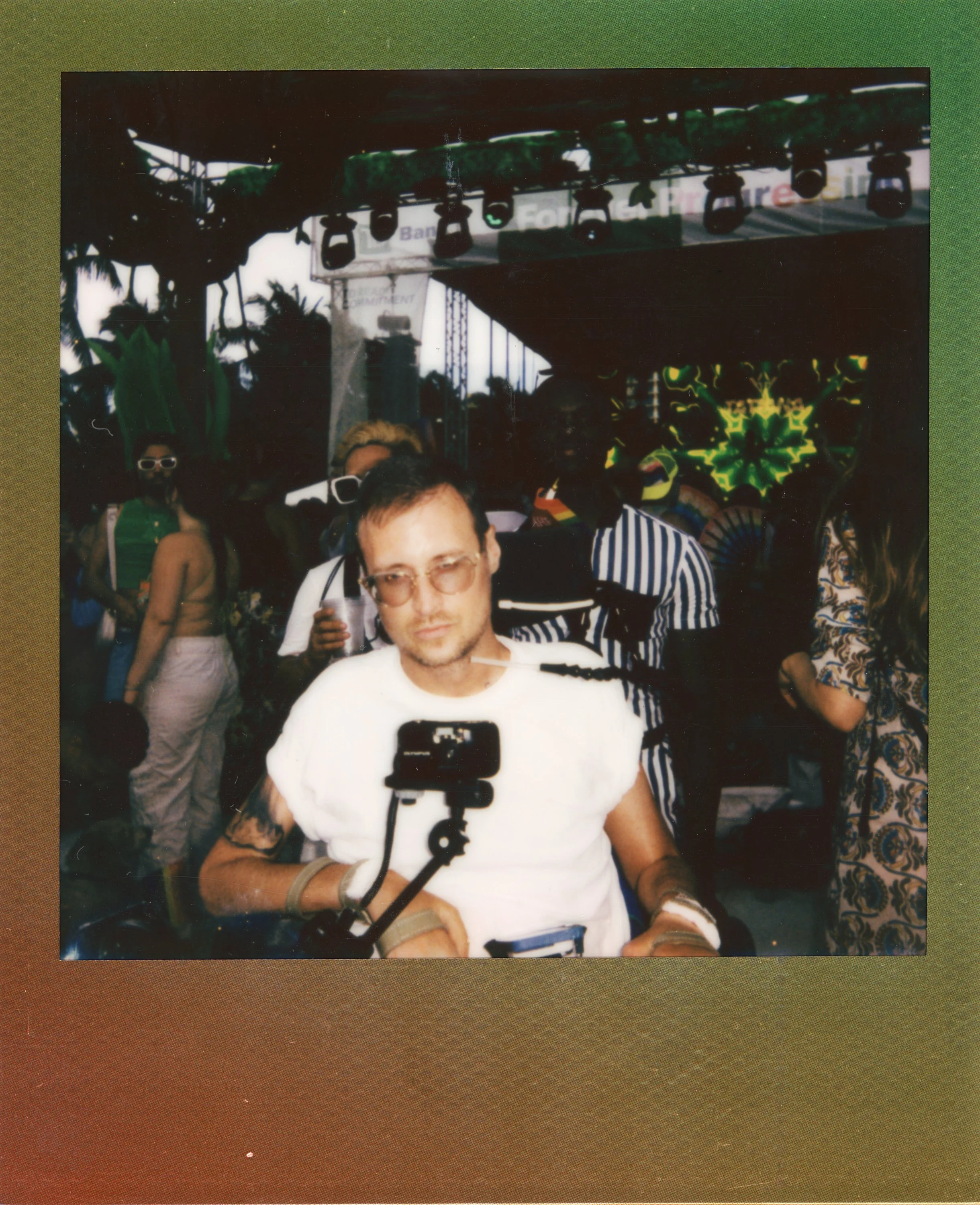 A man wearing sunglasses and a white shirt taking a selfie at a lively outdoor event or festival, with colorful lights and other attendees in the background.
