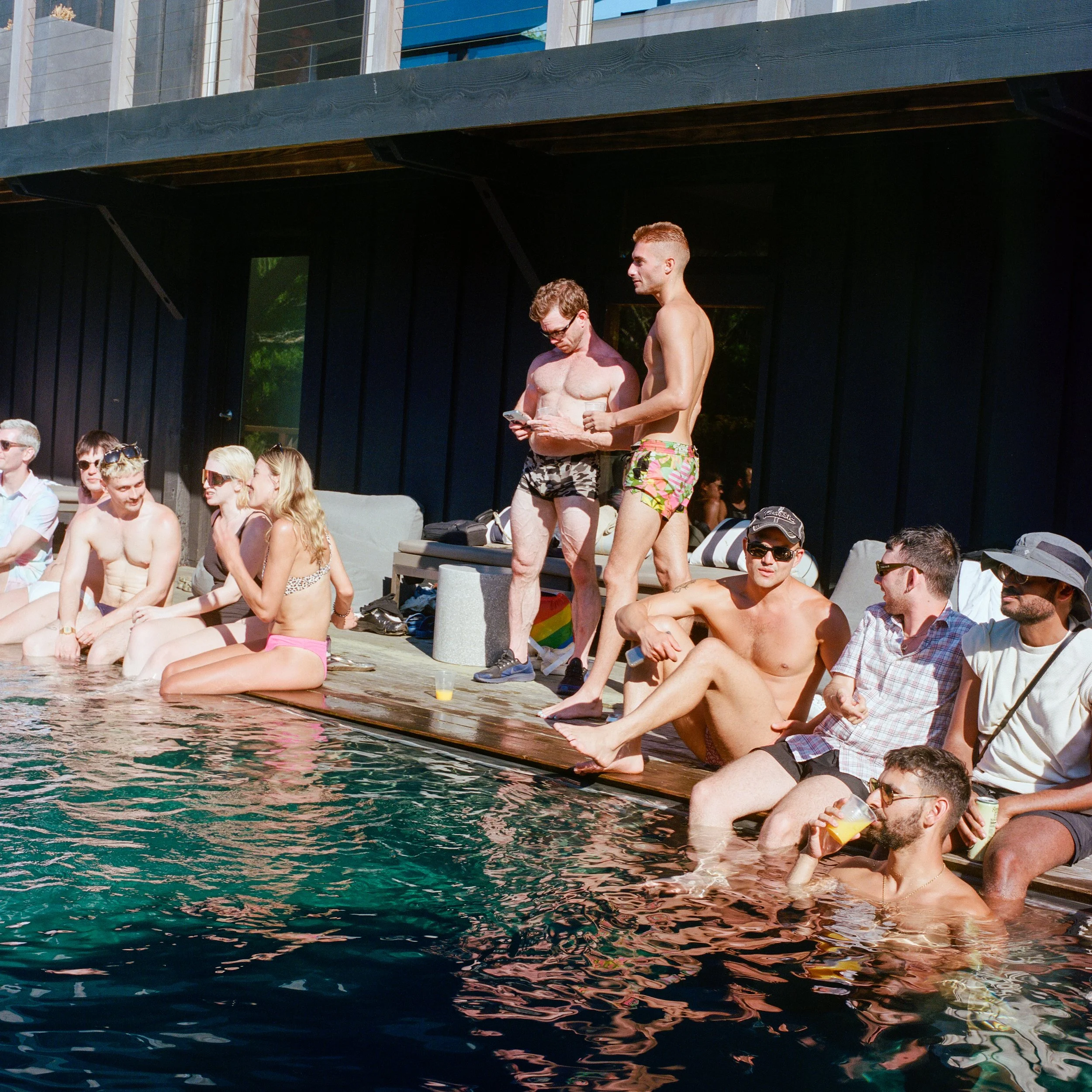 People gather by an outdoor poolside during a sunny day, some sitting on the edge with their feet in the water, others standing or sitting on lounge furniture, wearing swimsuits, sunglasses, and hats.