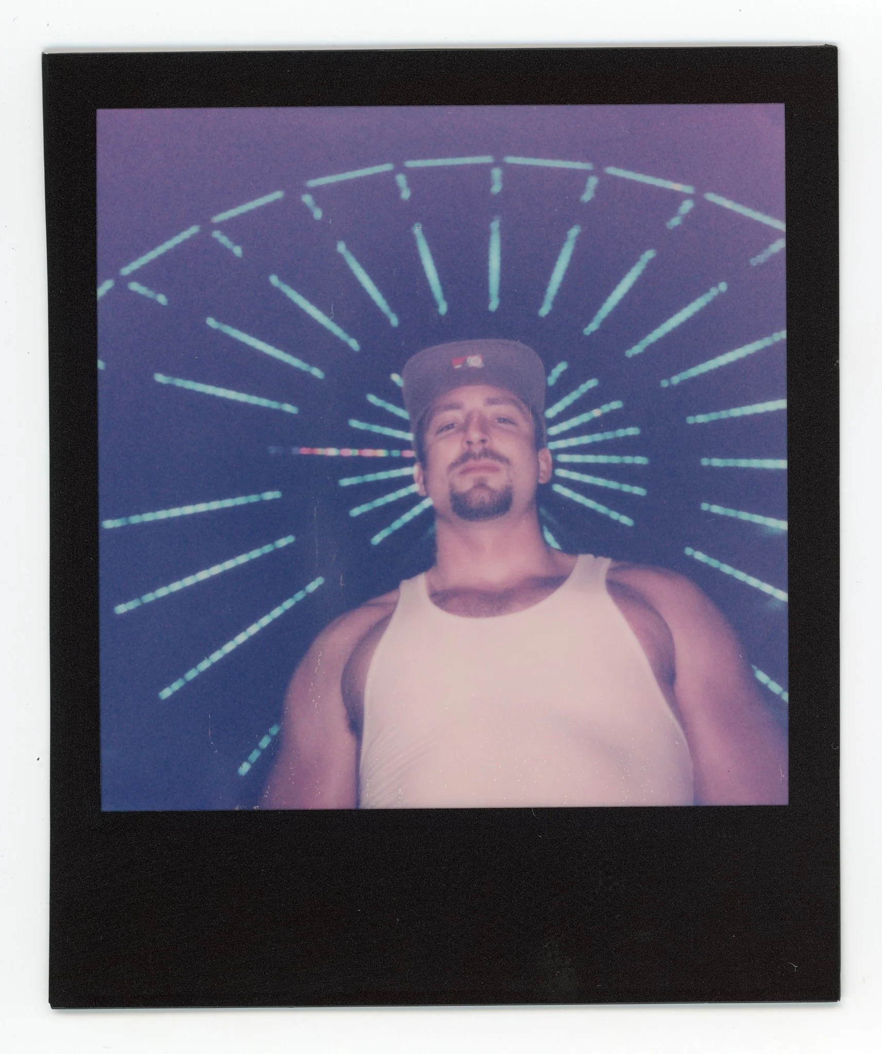 A man lying on his back in front of a star-shaped neon light display, wearing a white tank top and a gray cap.