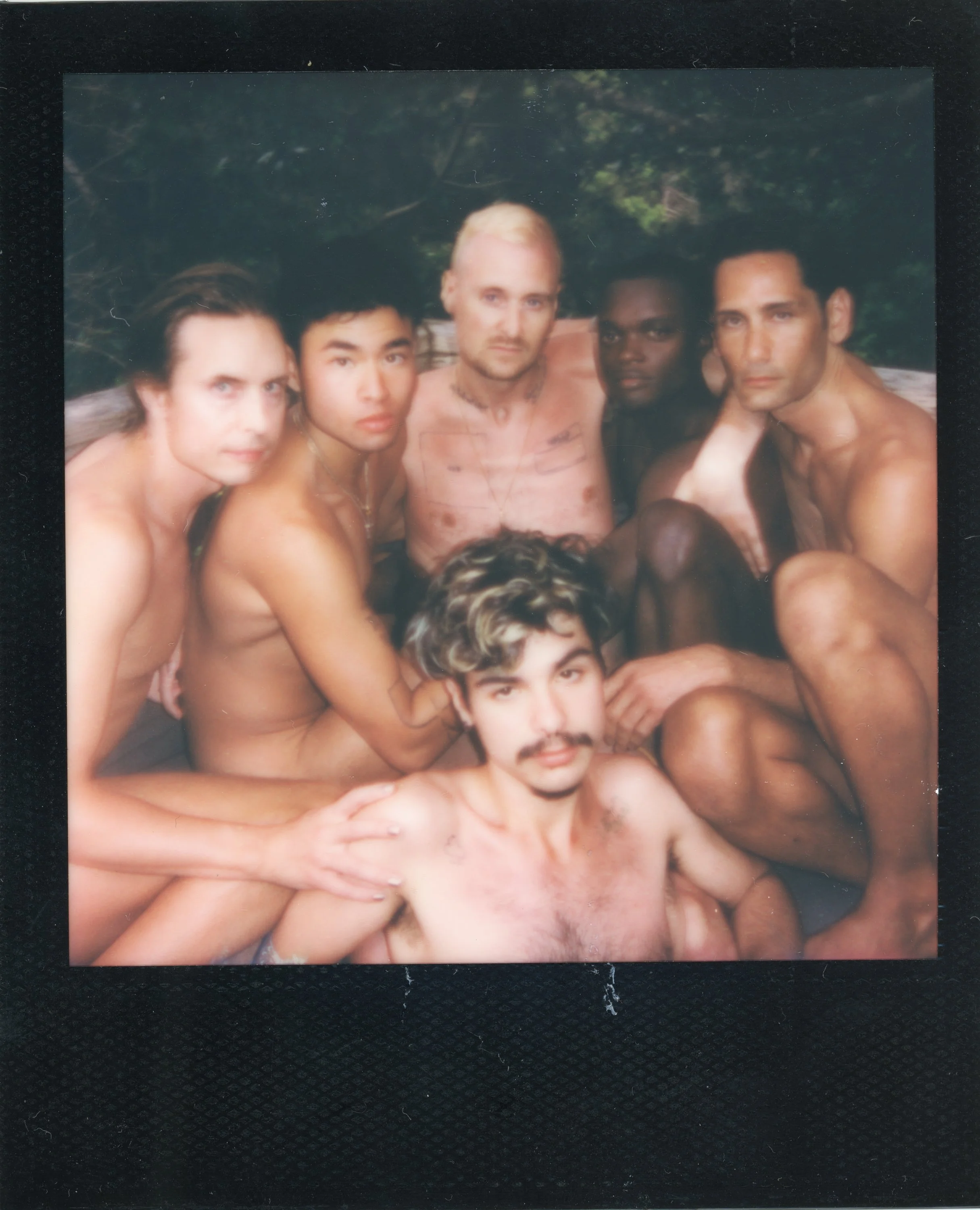 Group of six shirtless men with diverse skin tones and varying hairstyles, sitting closely together outdoors with green foliage in the background.
