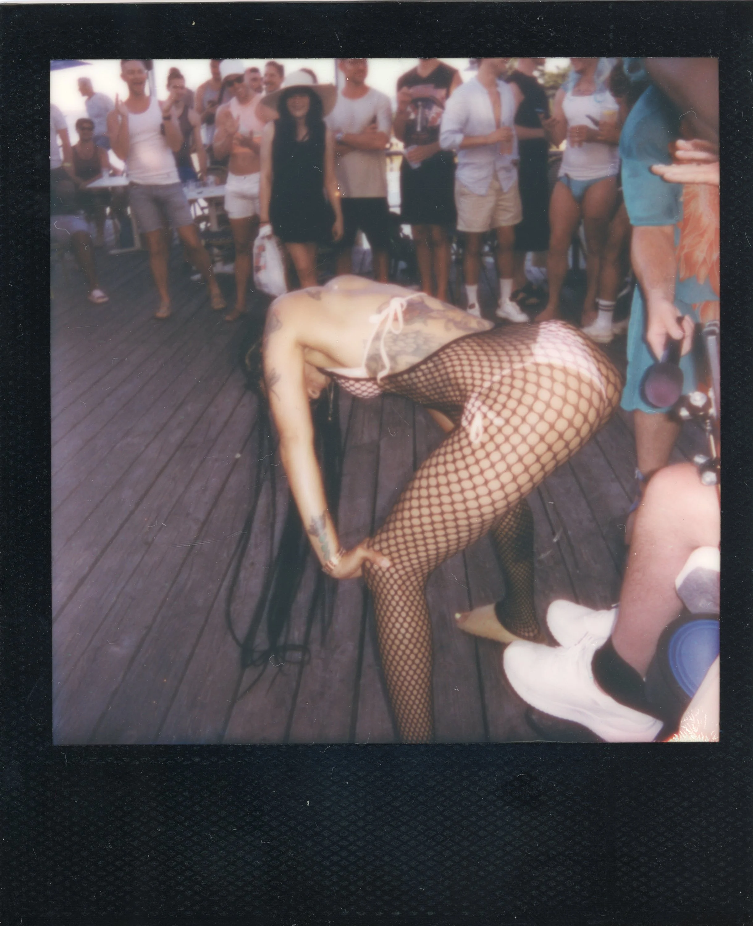 A woman with tattoos and long dark hair bending forward on a wooden deck, wearing fishnet stockings and a swimsuit, surrounded by a group of people watching her perform or participate in an activity.