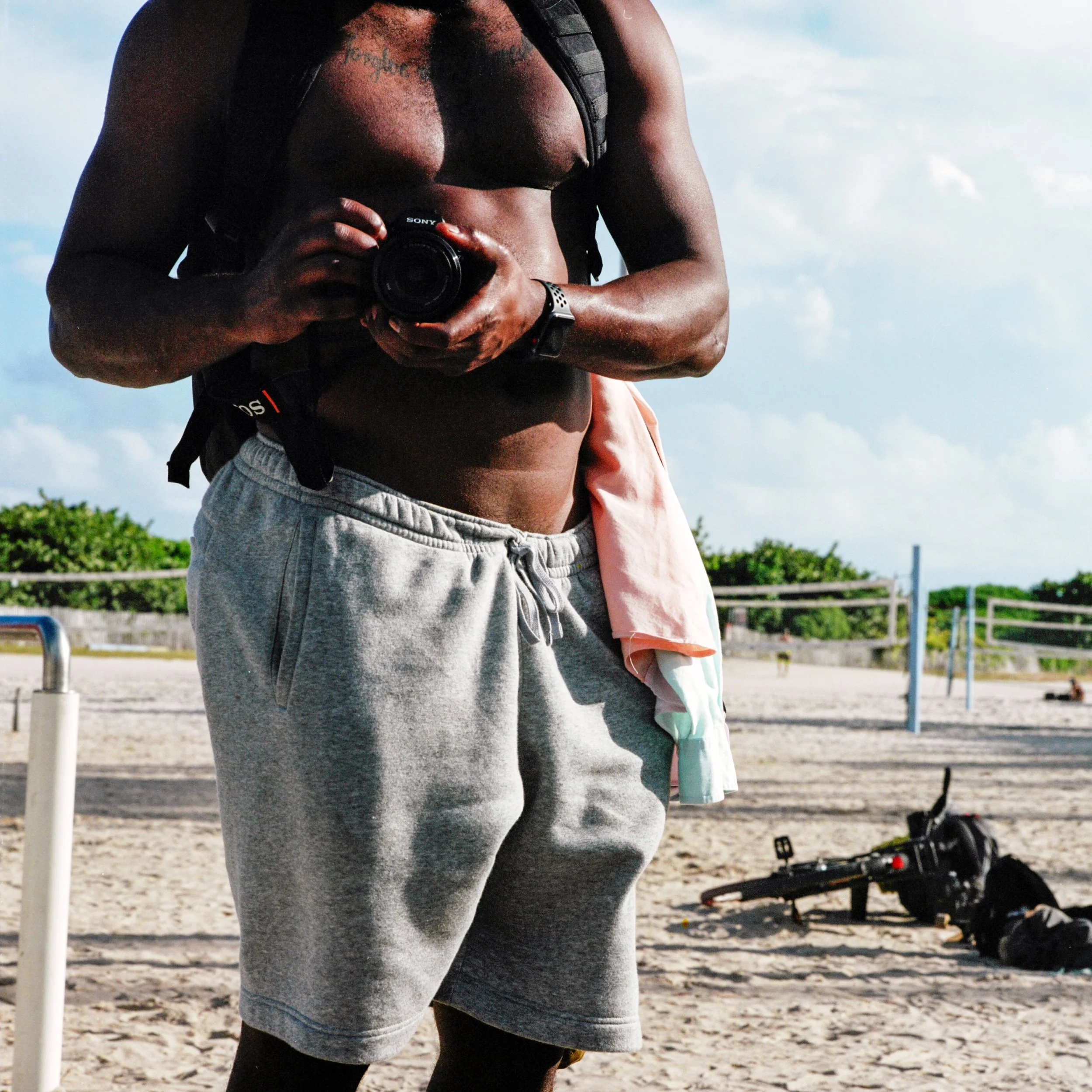 A shirtless man in gray shorts holding a camera at the beach with volleyball nets and a bag in the background.