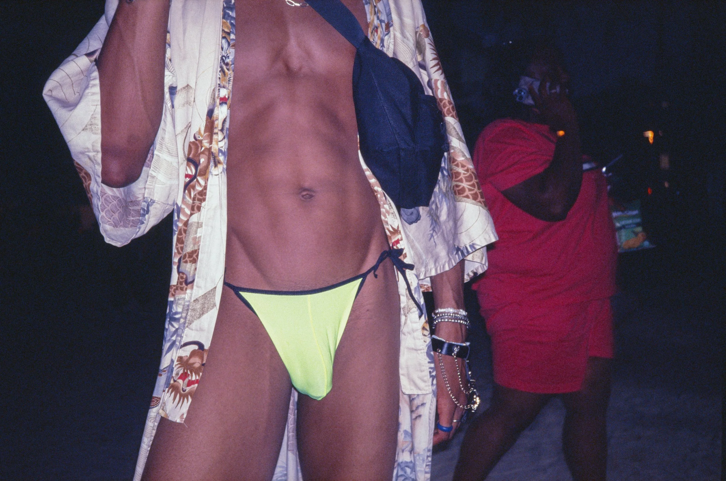 Person wearing a patterned shirt, yellow bikini bottom, and black accessories standing next to someone in a red shirt near the beach at night.