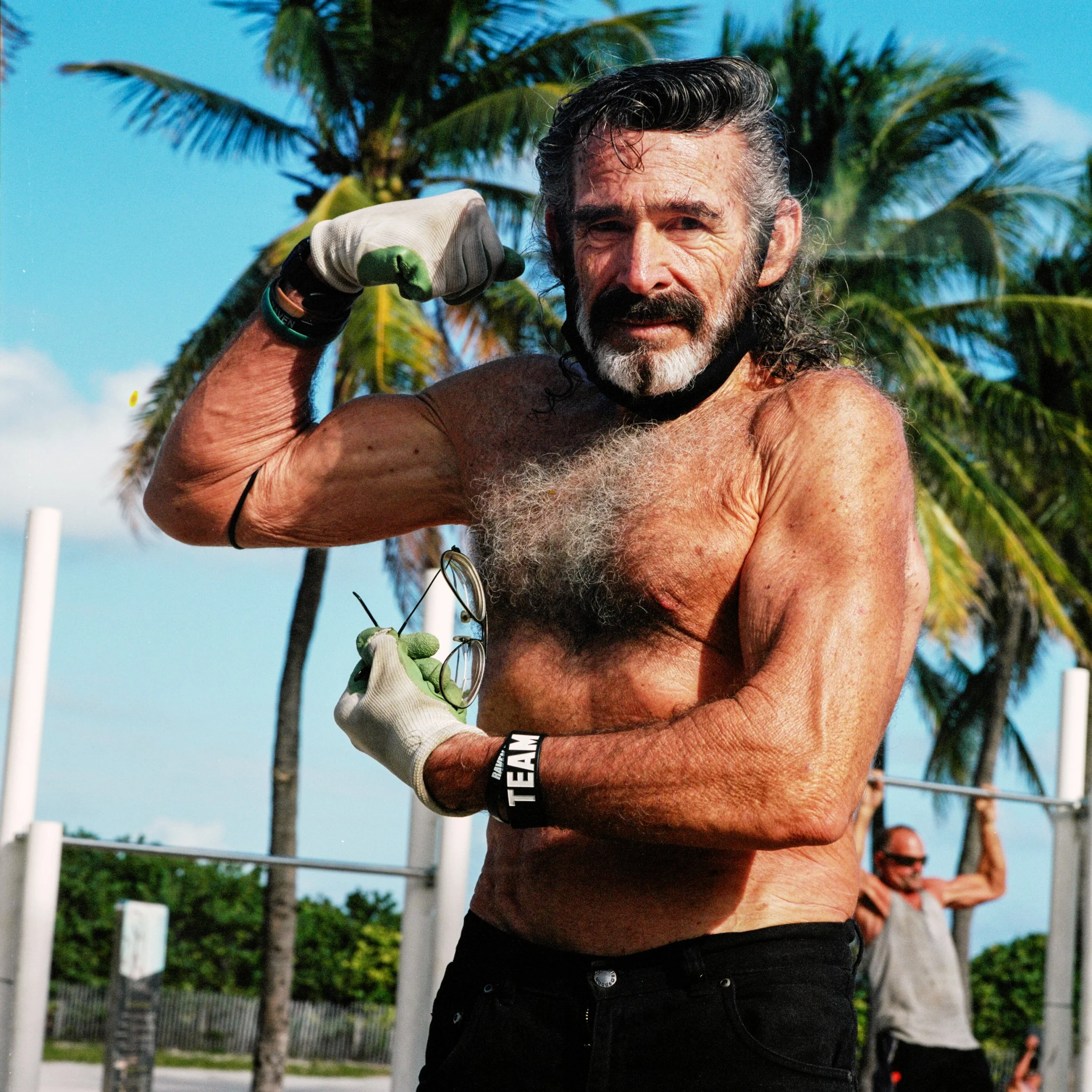 A shirtless older man with gray hair and a beard flexes his bicep while holding glasses and wearing workout gloves outdoors with palm trees in the background.