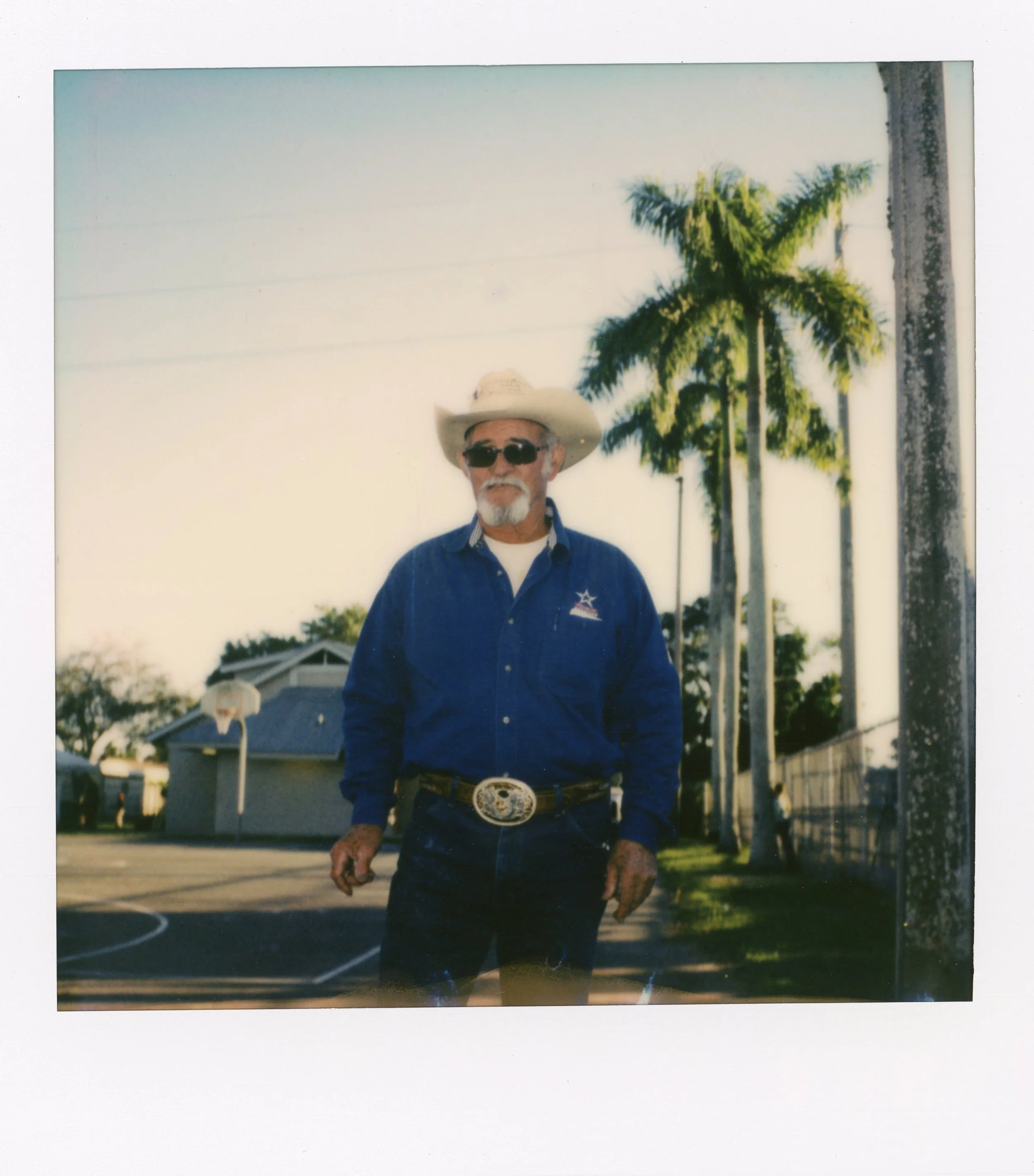 An older man with a white beard and sunglasses standing outdoors on a basketball court, wearing a cowboy hat, blue shirt, and belt with a large buckle, with palm trees and a house in the background.