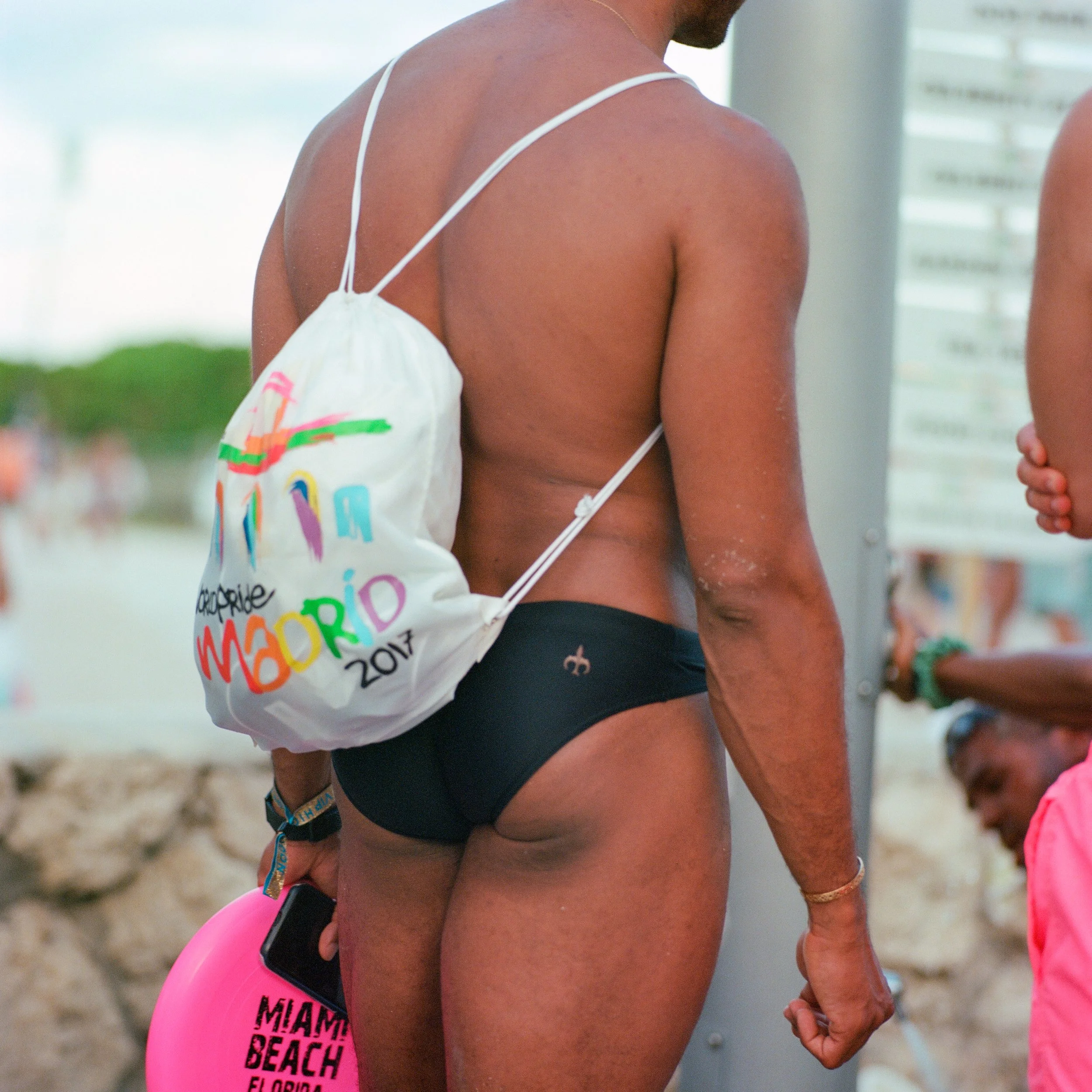 Person wearing black swim briefs and carrying a white drawstring bag with colorful writing at the beach