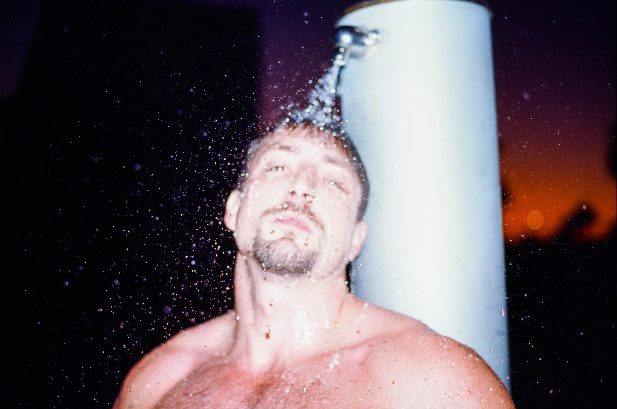 A man getting a cold shower with water streaming down his face and chest, with a dark background and orange hue in the sky.