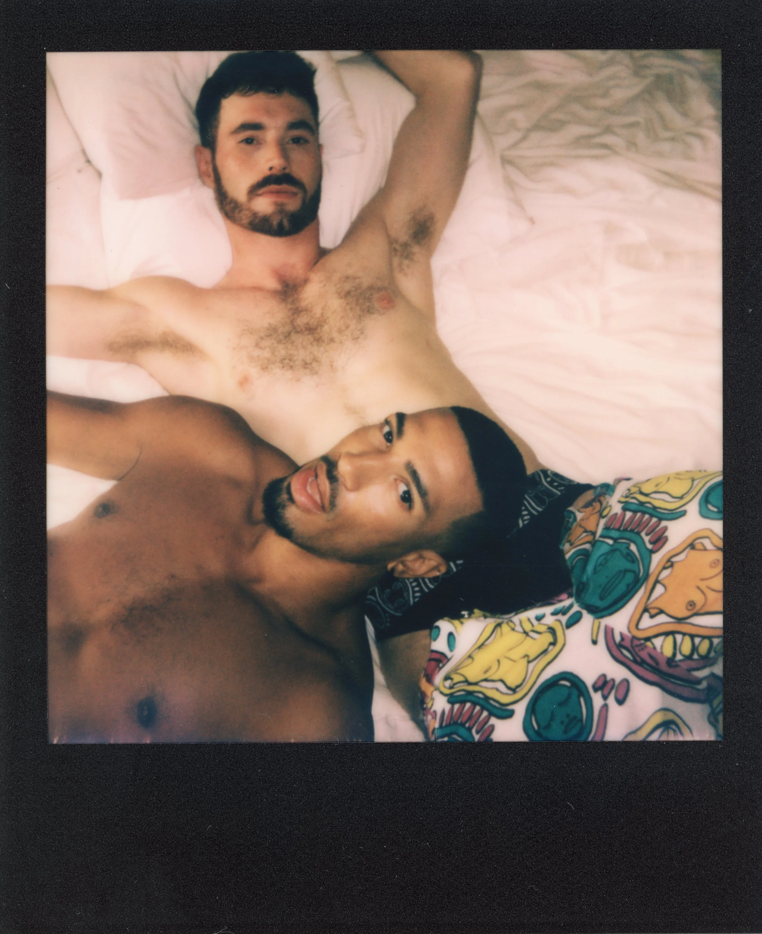 Two shirtless men lying in bed, one with light skin and dark hair, the other with tan skin and dark hair, taking a selfie. One man is smiling and sticking his tongue out, while the other has a more neutral expression. They are on pink bedsheets, and