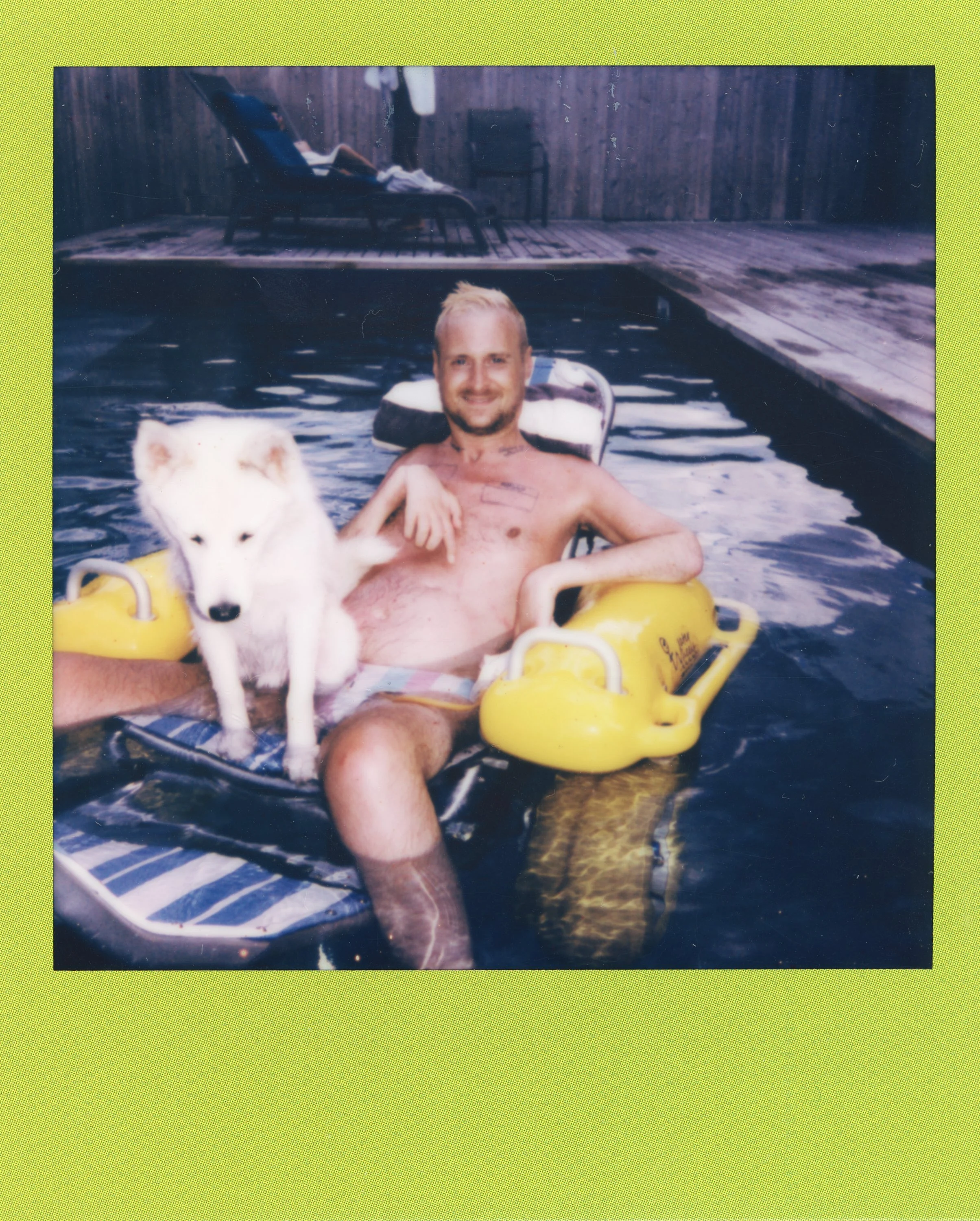 A man enjoying a swim in a backyard pool with a Siberian Husky dog sitting on his lap, sitting on a yellow float, with a wooden deck and outdoor furniture in the background.