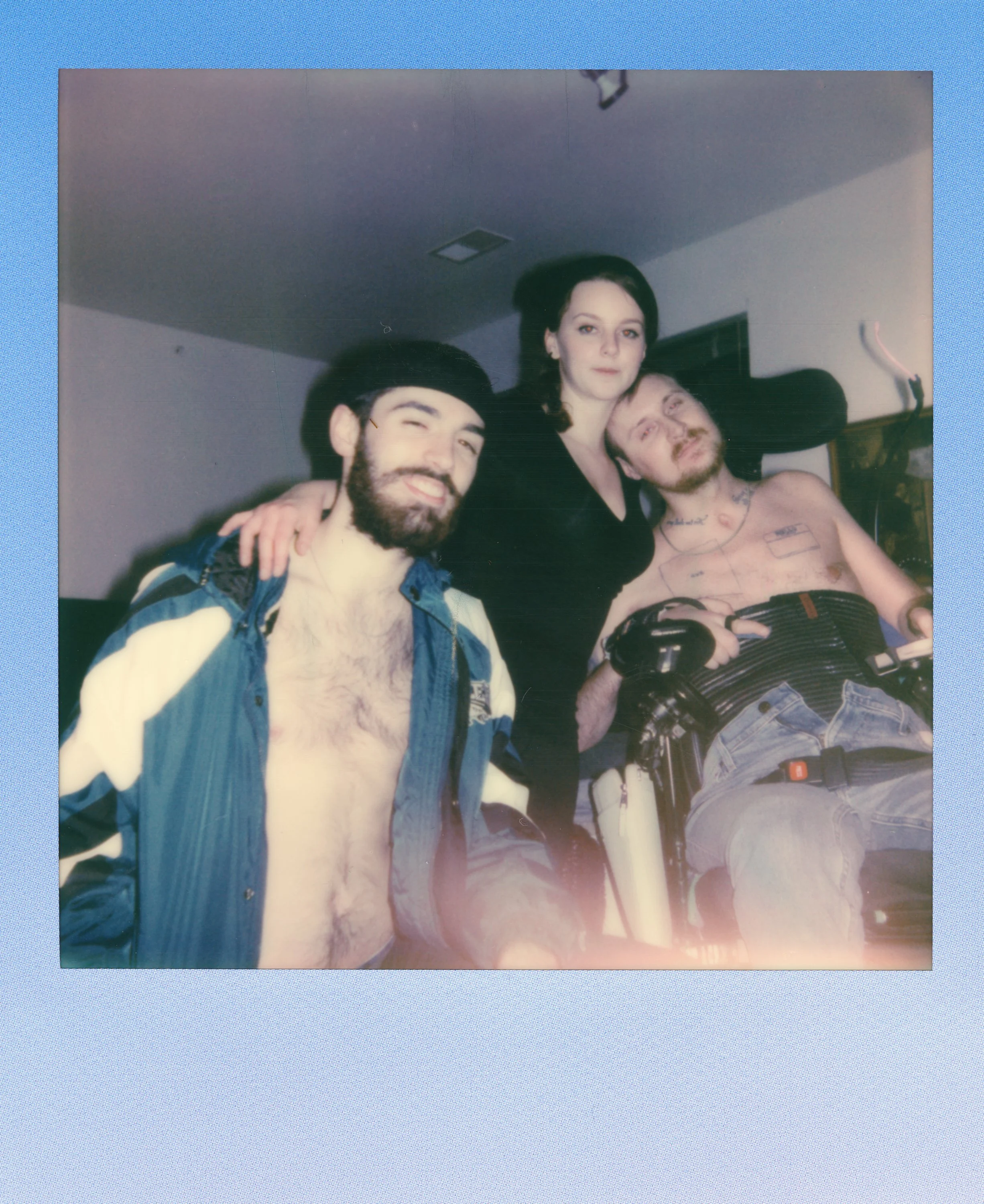 Three young adults posing together indoors, one shirtless man with tattoos and wheelchair, woman in black dress with long dark hair, smiling man with a beard and open jacket, casual setting.
