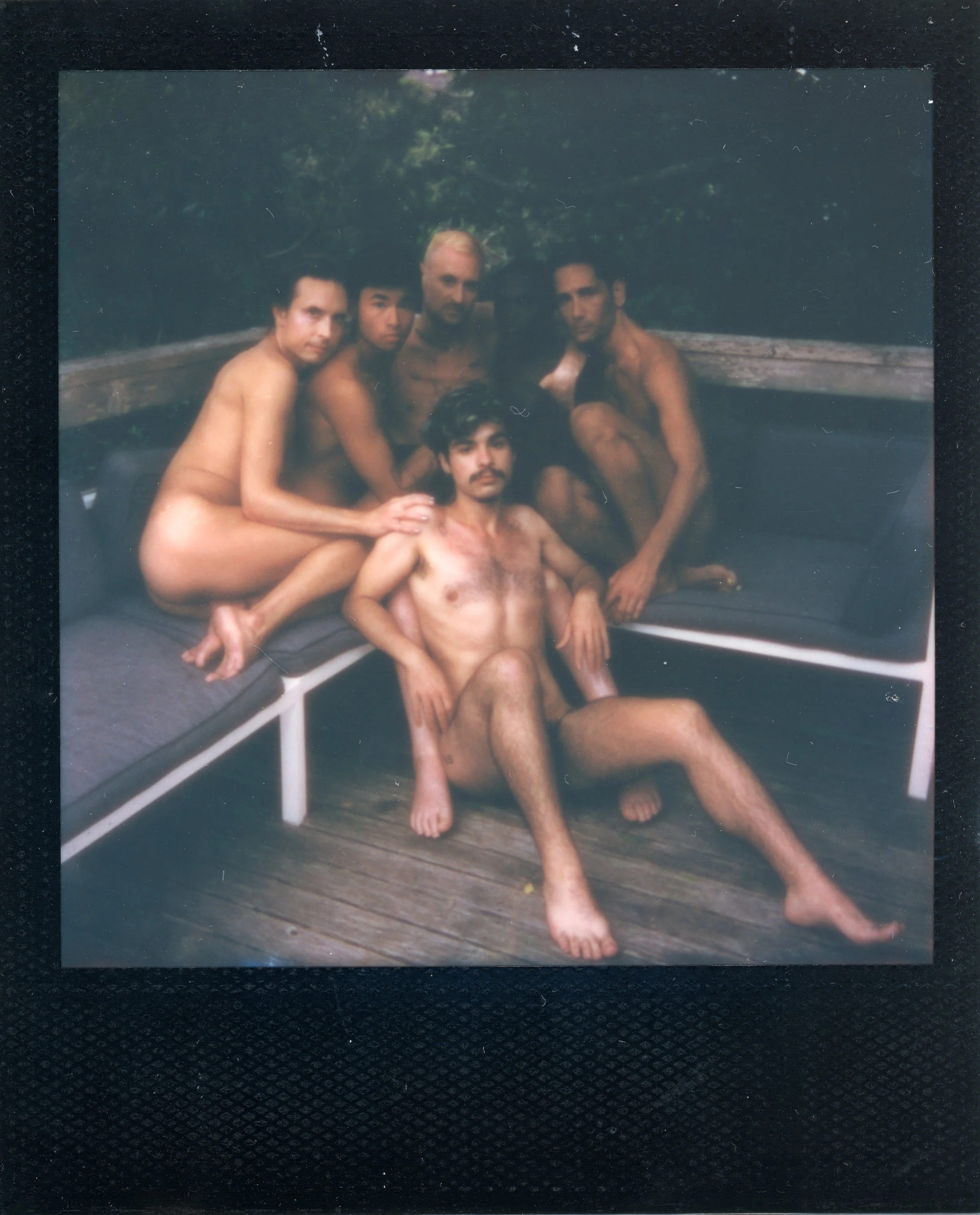 Group of five nude men sitting and lounging outdoors on a wooden deck with greenery in the background.