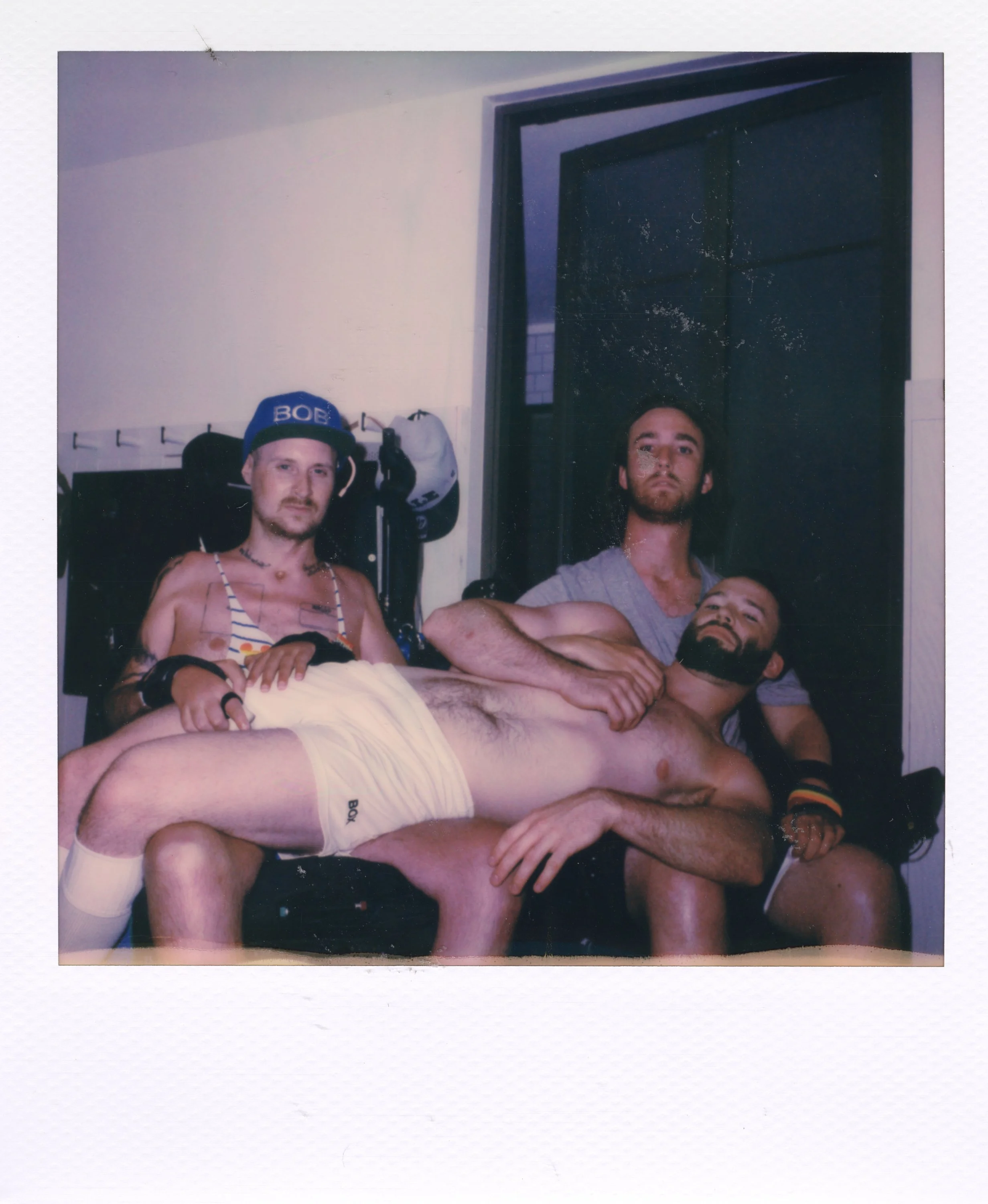 Three men are posing together in a room, with one lying across the laps of the other two. The man lying down is shirtless, wearing yellow underwear, and has a beard. The man in the middle has long dark hair, a beard, and is wearing a gray t-shirt. Th