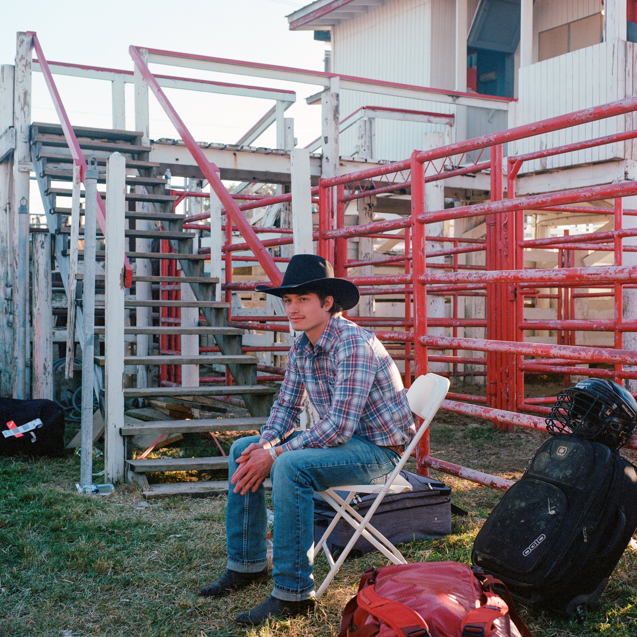 Young man in plaid shirt and black cowboy hat sitting on a folding chair beside rodeo fencing and gear.