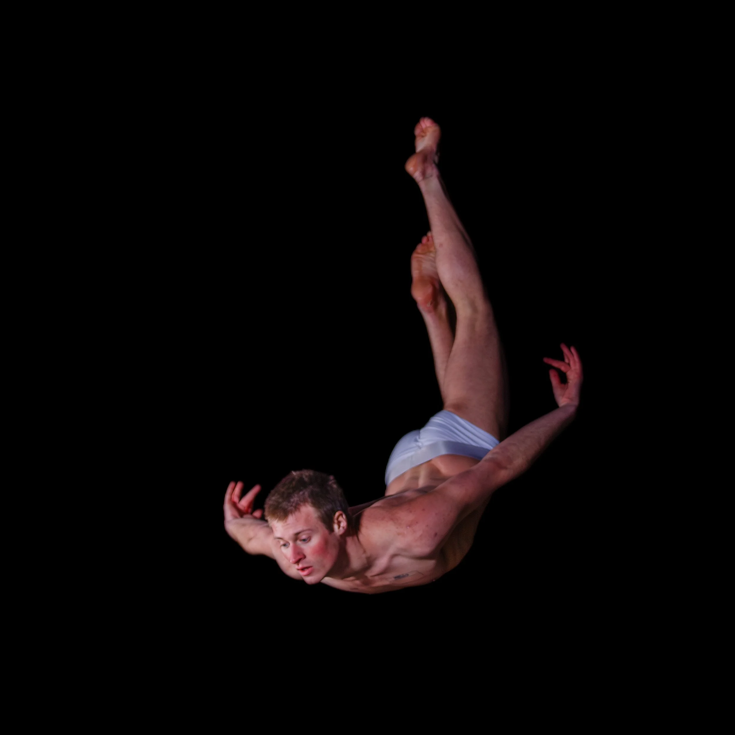 A male dancer in white shorts performing a mid-air leap against a black background.