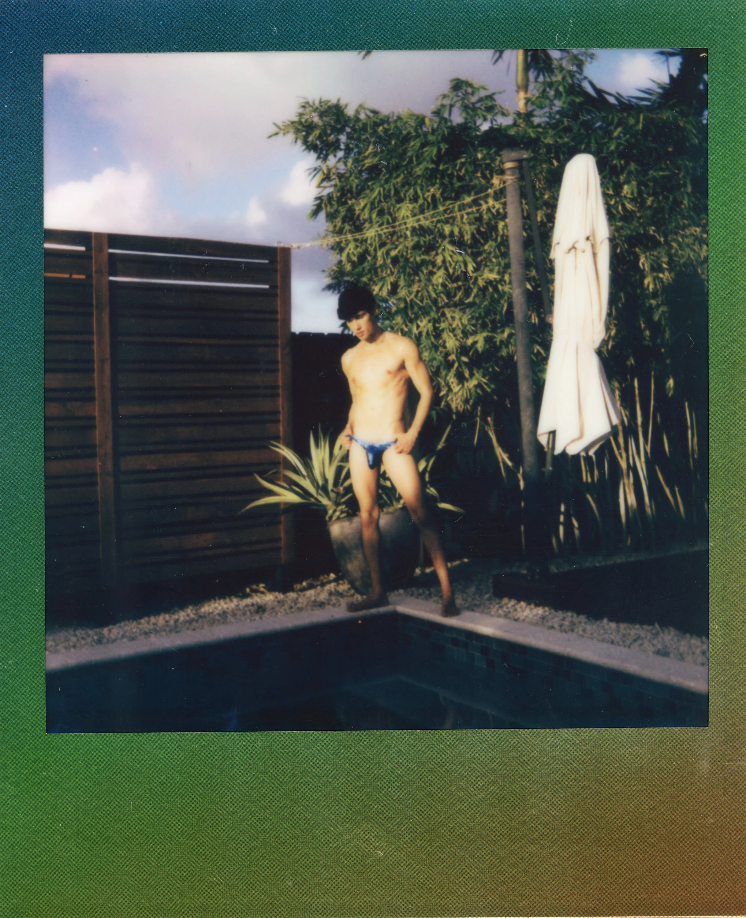 A shirtless young man wearing swim trunks standing by a swimming pool with a wooden fence, large green plants, and an umbrella in the background.