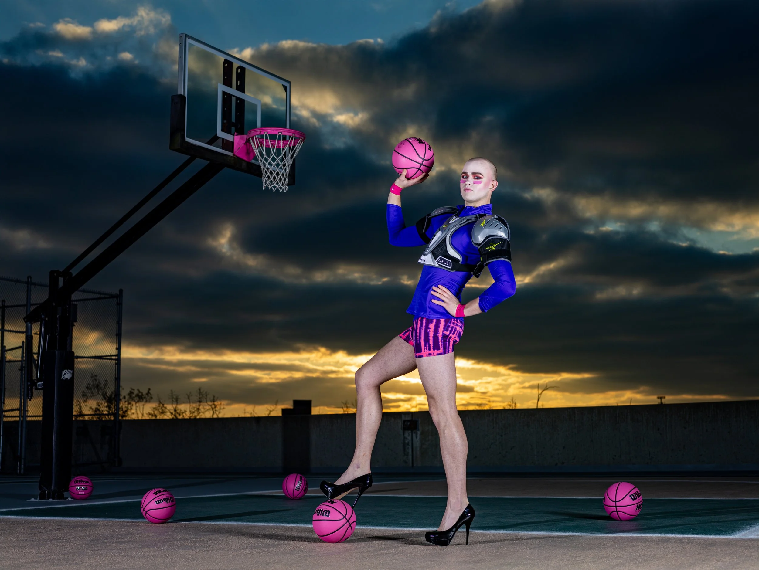 A person dressed in athletic and futuristic clothing standing on an outdoor basketball court at sunset, holding a pink basketball in one hand, with multiple pink basketballs on the ground, and a basketball hoop in the background.