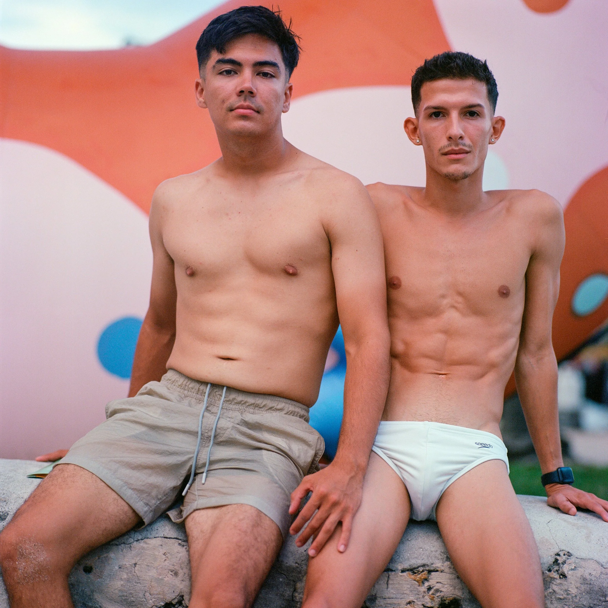 Two shirtless men sitting on a concrete ledge in front of a colorful abstract background, wearing underwear and shorts.