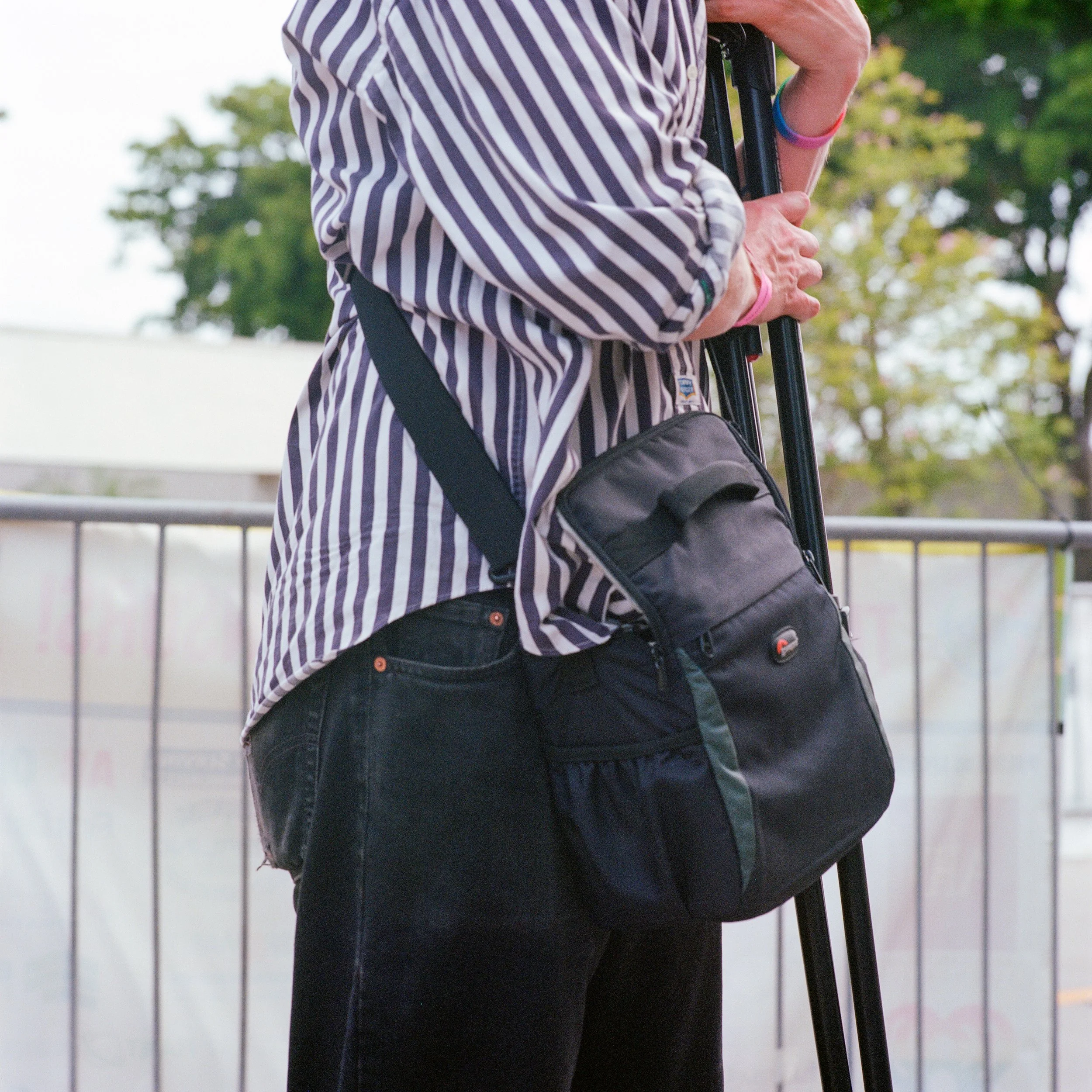 Person holding a tripod and carrying a black bag, wearing a striped shirt and black pants outdoors.