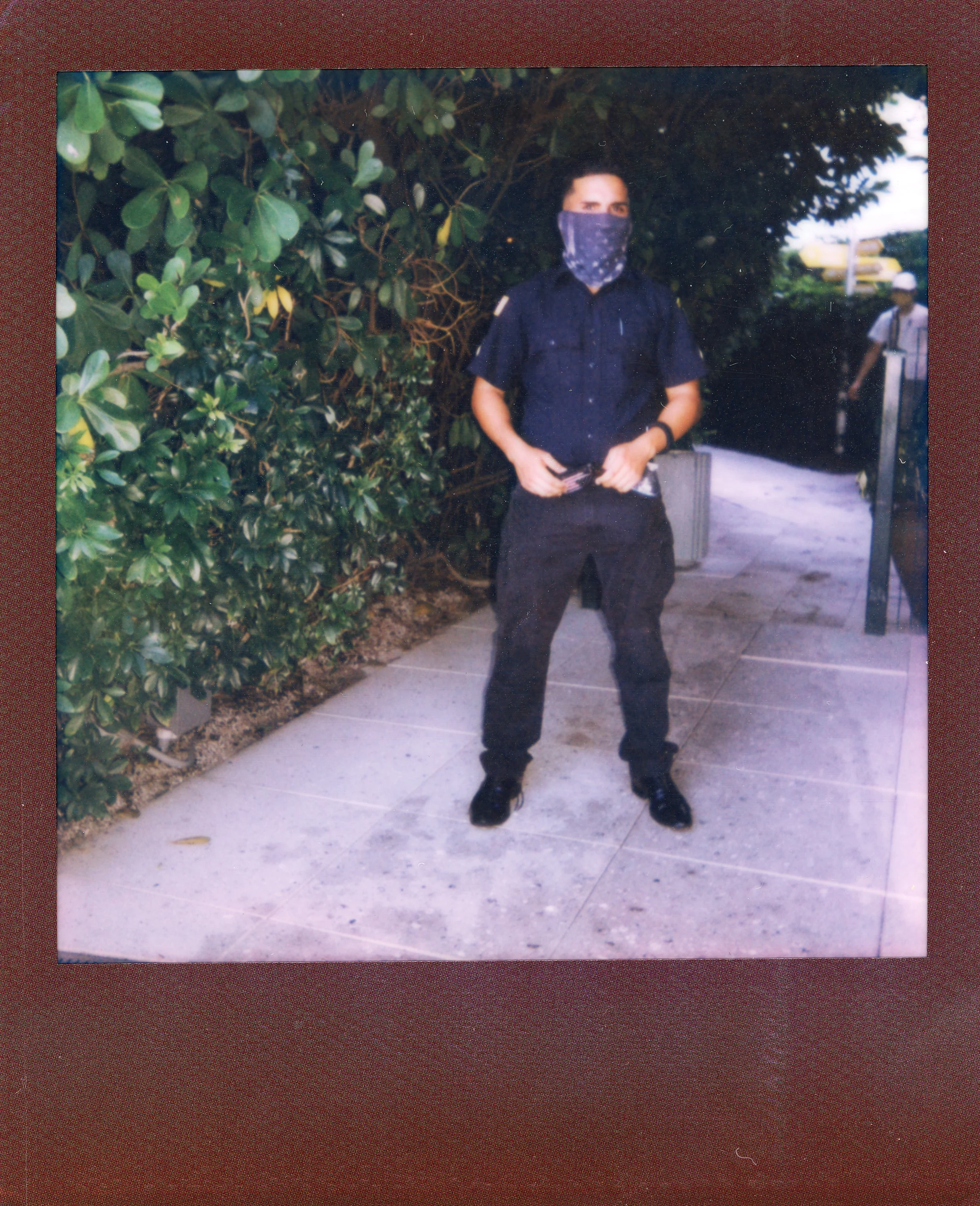 A man wearing a dark uniform and a face mask stands outdoors on a sidewalk next to green bushes, with another person in the background.