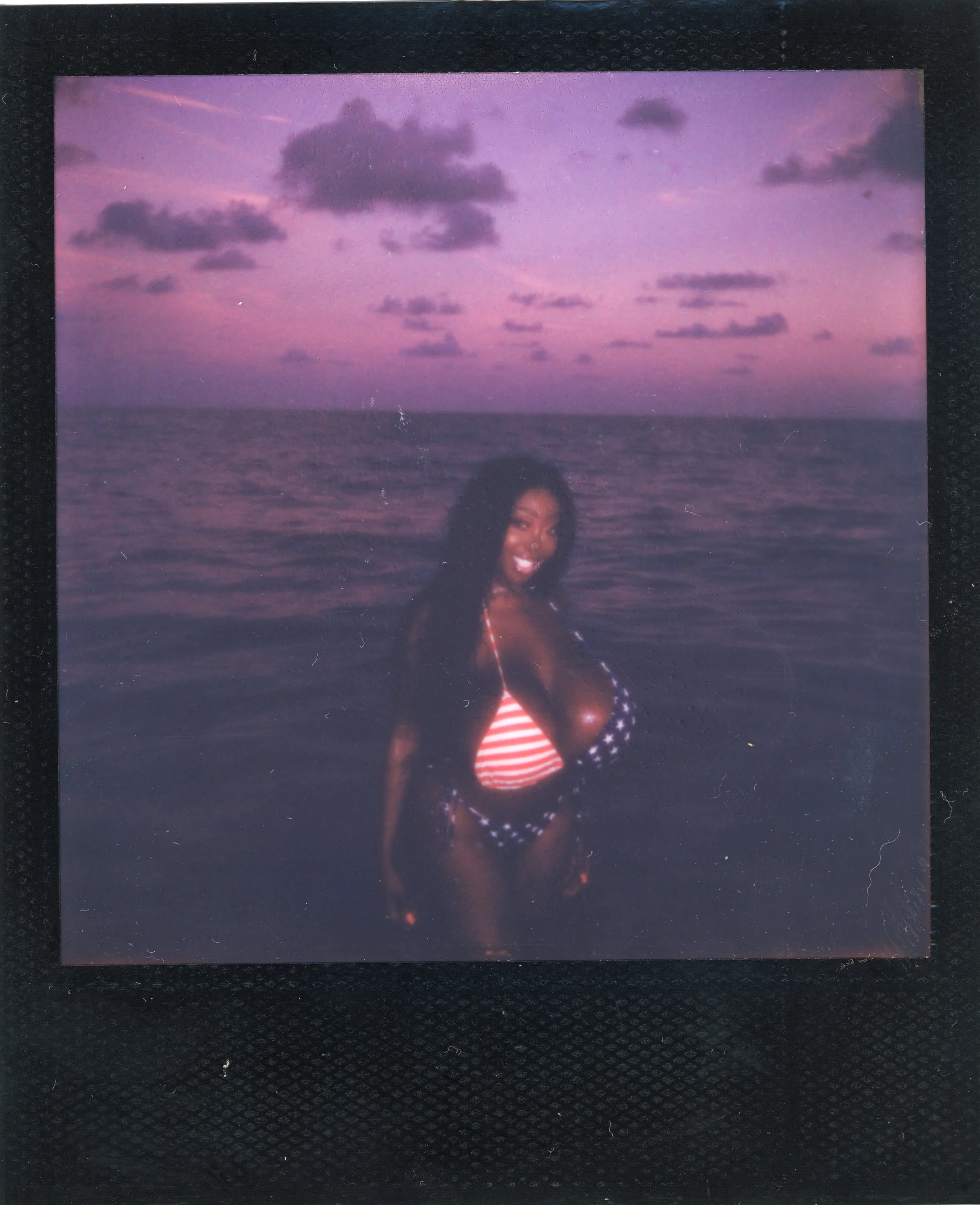 A woman in a swimsuit with an American flag pattern standing in the ocean at sunset, smiling and looking back at the camera.