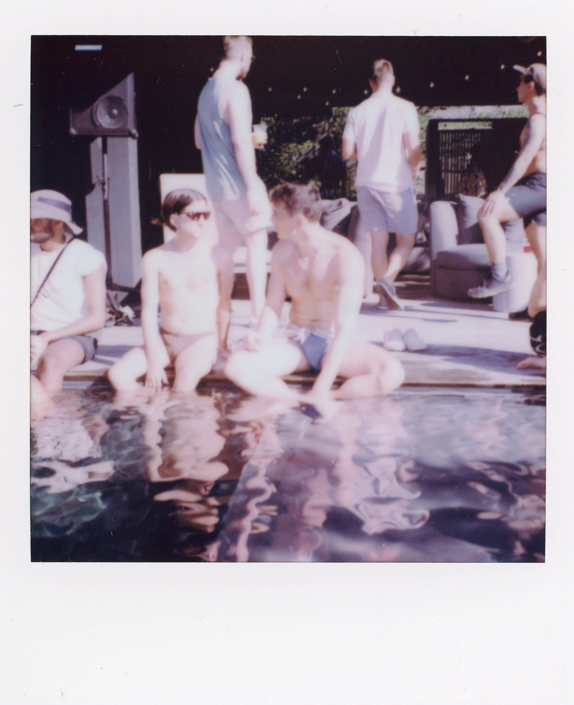 Group of young people at a poolside, some sitting and some standing, with a pool in the foreground and outdoor furniture in the background, during sunny weather.