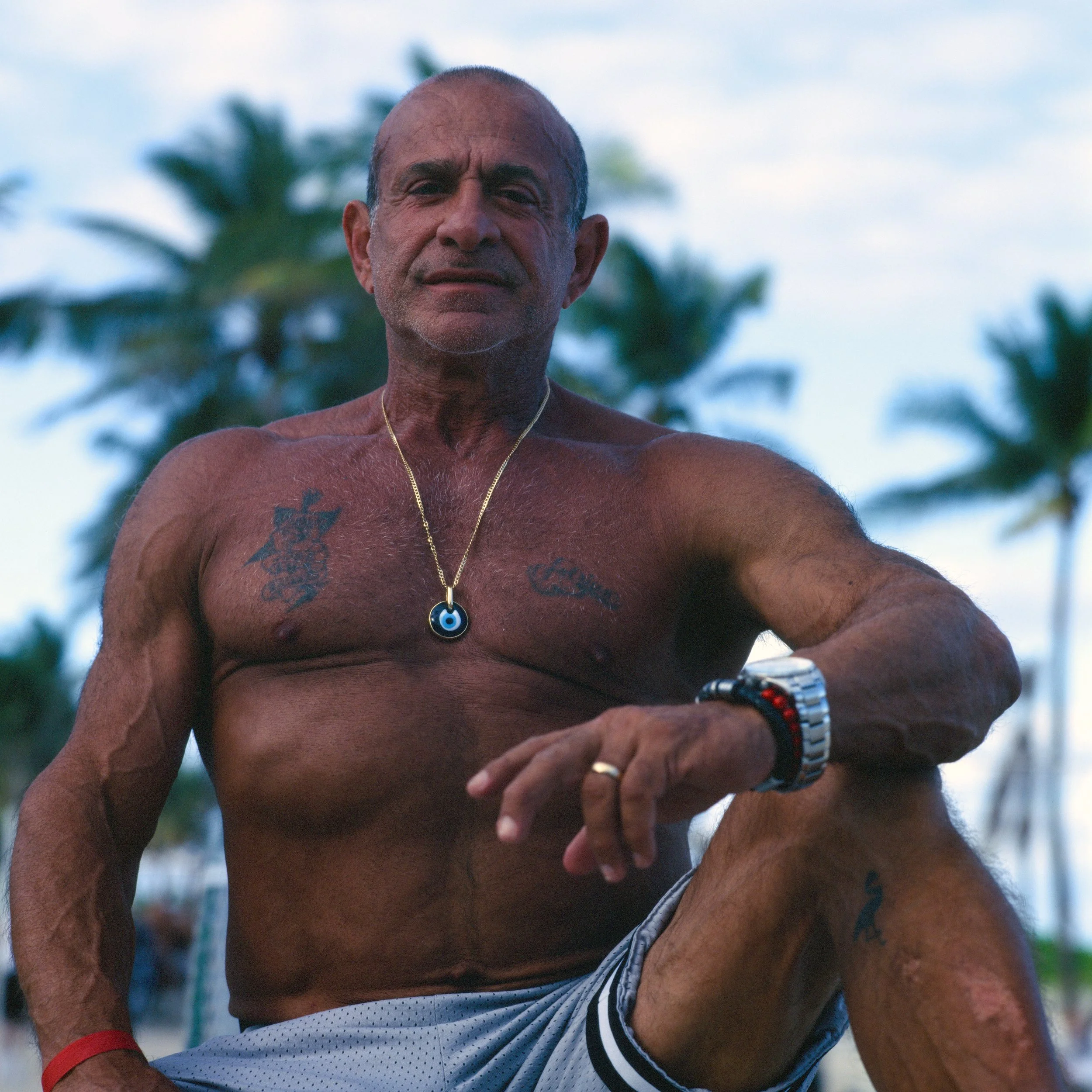 A muscular, shirtless older man with tattoos, wearing a gold necklace with an evil eye pendant, a wristwatch, and a bracelet, is posing outdoors with palm trees in the background.
