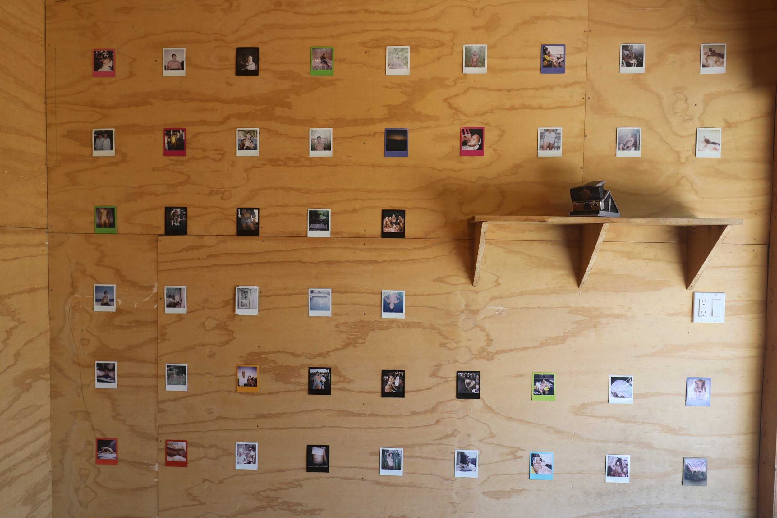 Wood-paneled wall decorated with multiple small, colorful-framed photographs arranged in rows, with a small wooden shelf holding a camera or projector, and an electrical outlet visible at the lower right.