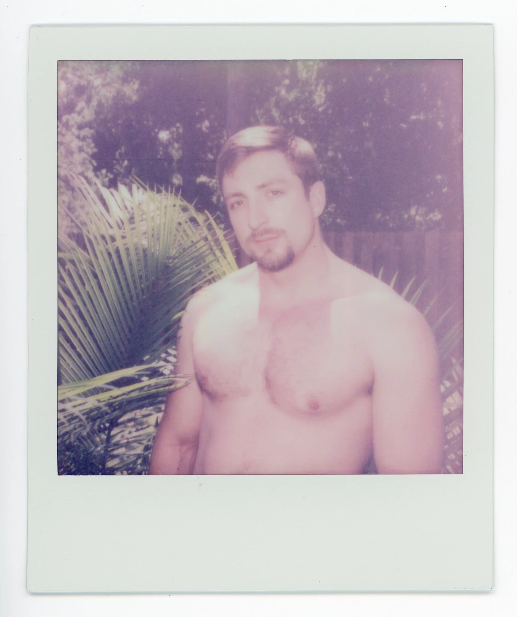 A shirtless man with short dark hair and a beard standing outdoors near a palm plant, with trees and a fence in the background.