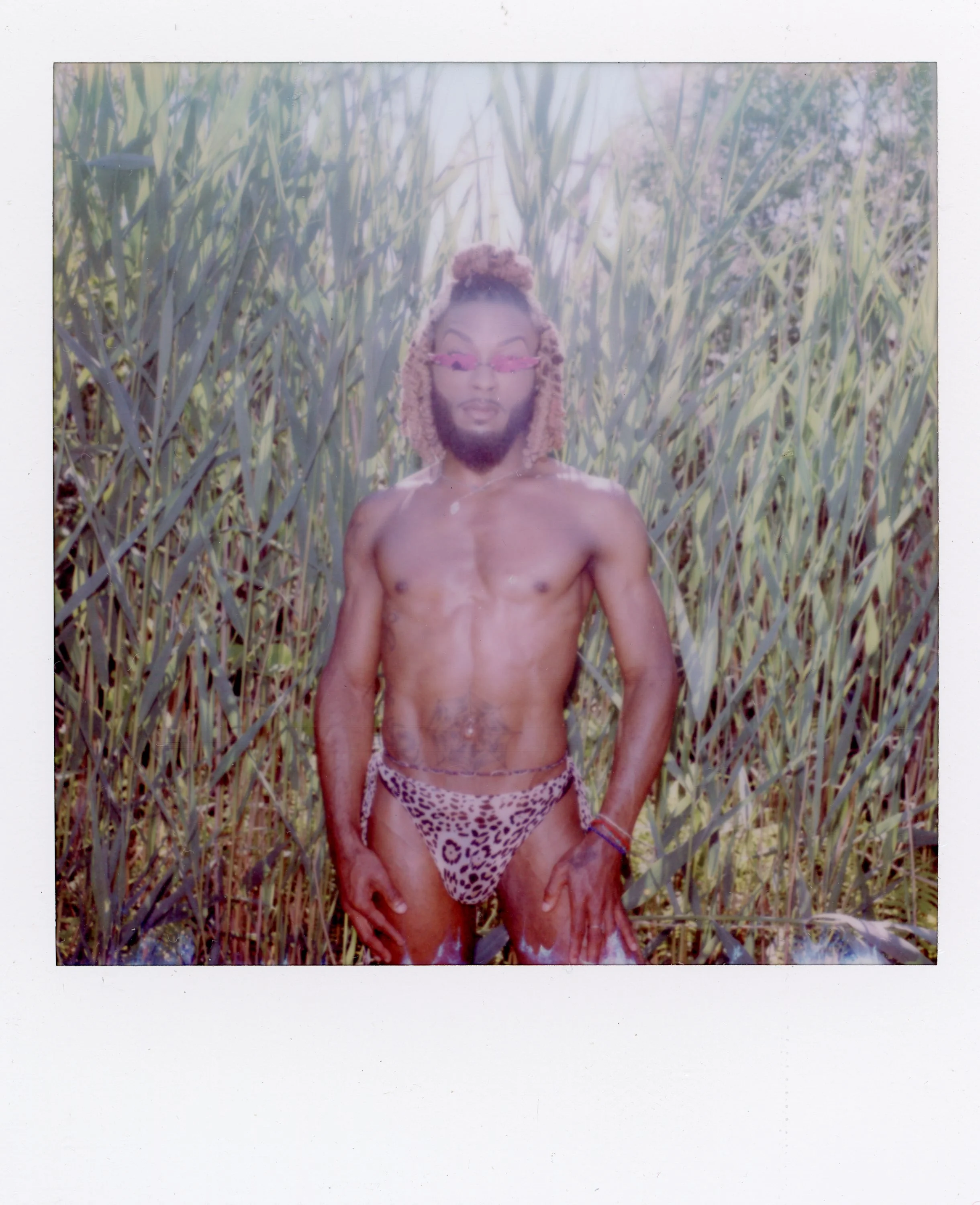 A shirtless man with dreadlocks and a beard standing in a field of tall grass, wearing animal print underwear and pink sunglasses.