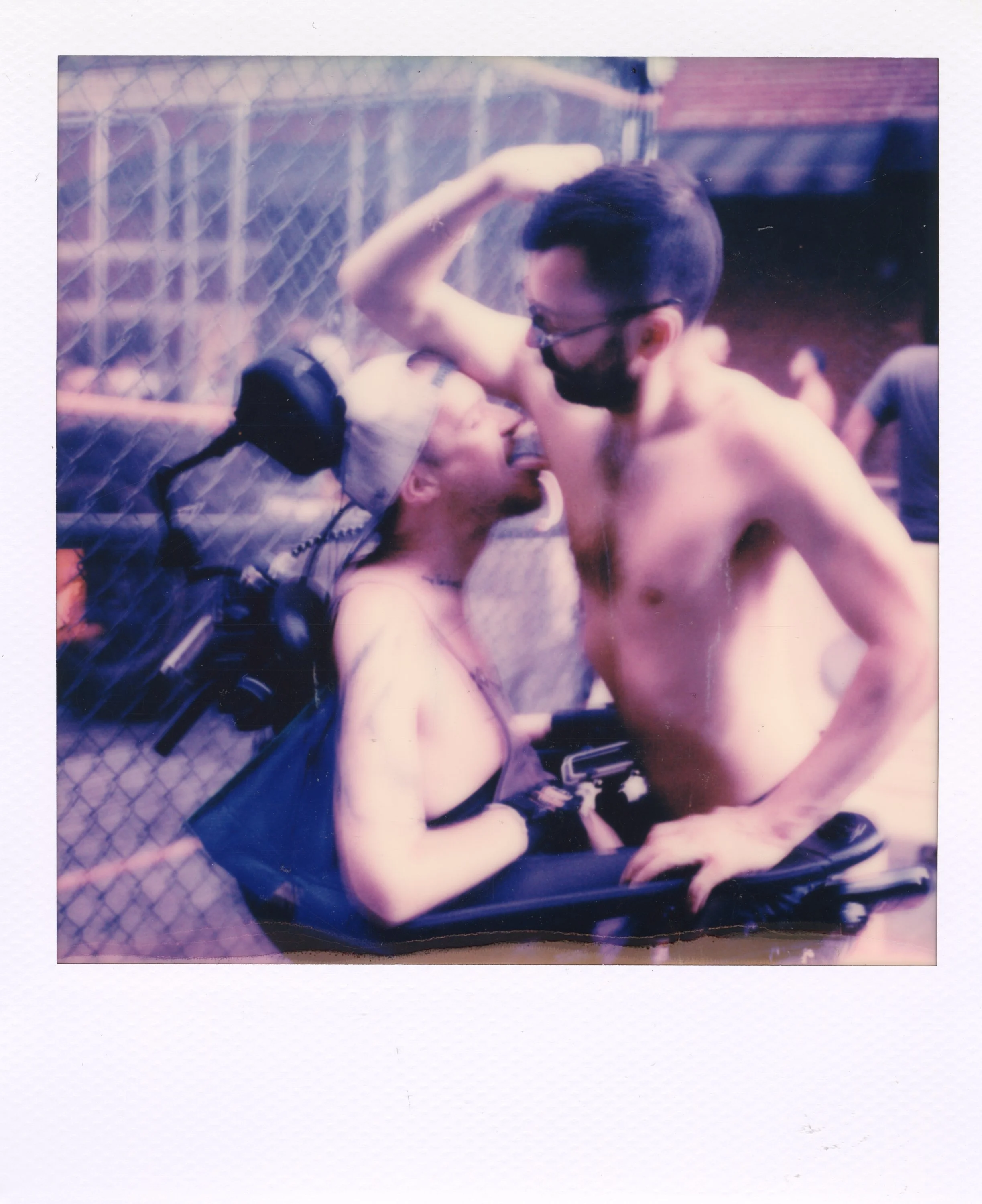 Two shirtless men are close to each other, with one leaning in and the other raising a fist, behind a chain-link fence. One man appears to be in a wheelchair wearing a cap, and the other is standing with a beard, glasses, and dark hair. They seem to