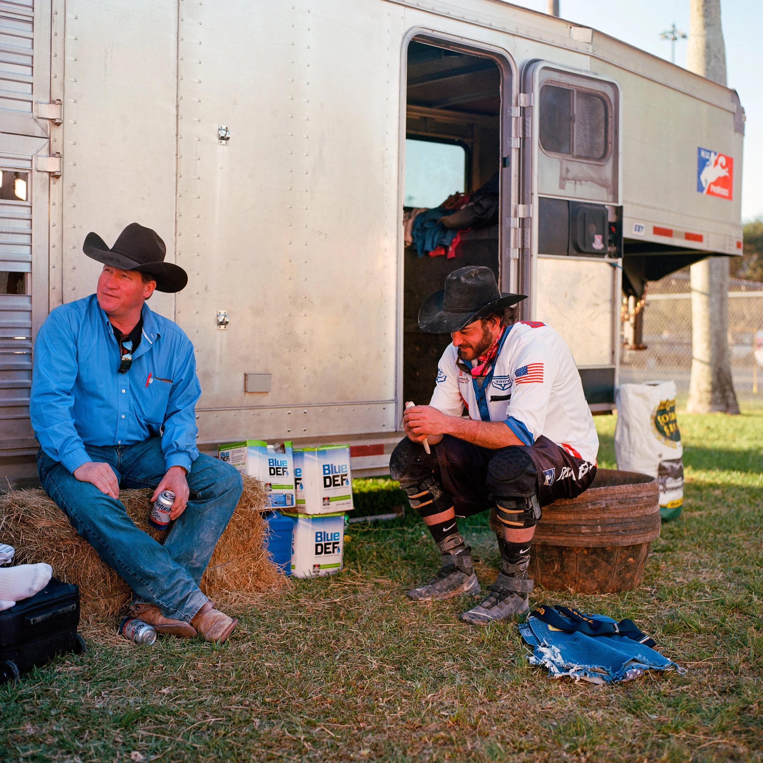 Two men in cowboy hats and Western-style clothing sitting outside near a silver trailer. One man in a blue shirt and jeans sitting on hay bale holding a beverage, the other in a white shirt with American flag patch, sitting on a circular metal contai