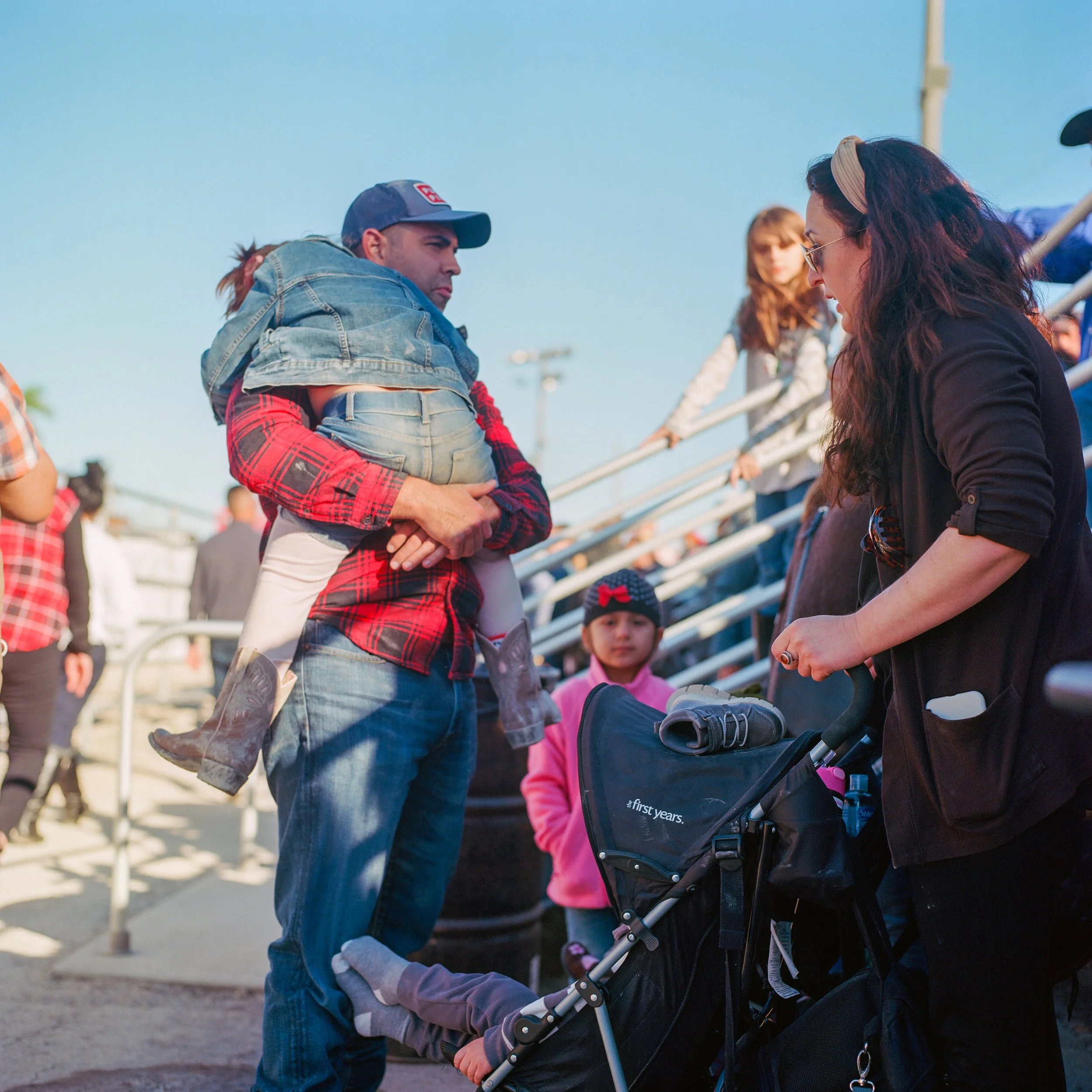 A man is holding a young girl on his shoulders, standing in a line at an outdoor event. A woman with sunglasses and a headband is leaning on a stroller, talking to the man. Several people are visible in the background, including children and adults,