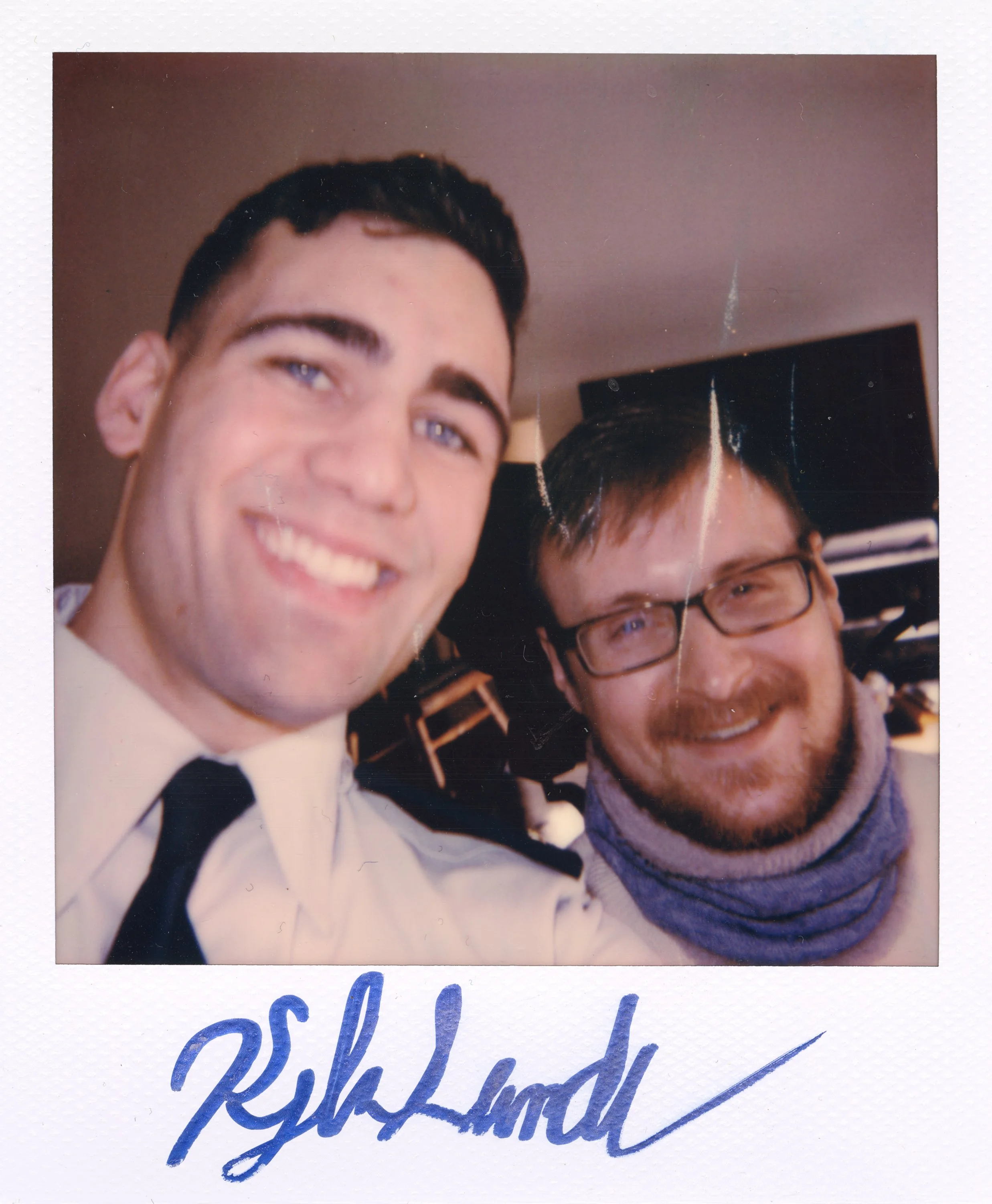 Two smiling men taking a selfie indoors; the man on the left is dressed in a white shirt and black tie, and the man on the right wears glasses and a scarf. There is a signature at the bottom of the photo.