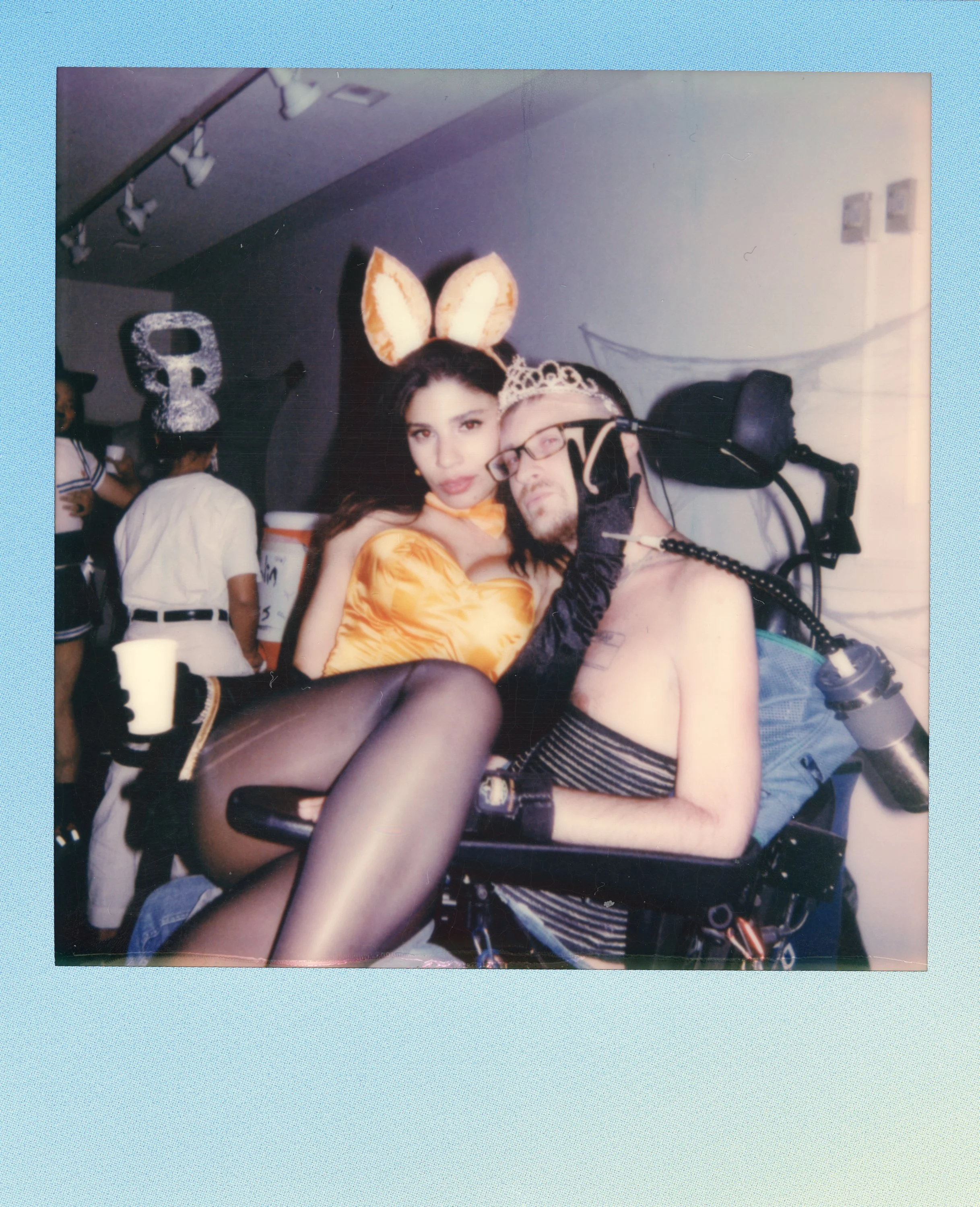 A woman dressed as a bunny and a man in a wheelchair with a tiara. The woman is holding a cup and wearing bunny ears, while the man is wearing a tiara and has a breathing apparatus. They are at a costume or themed event.