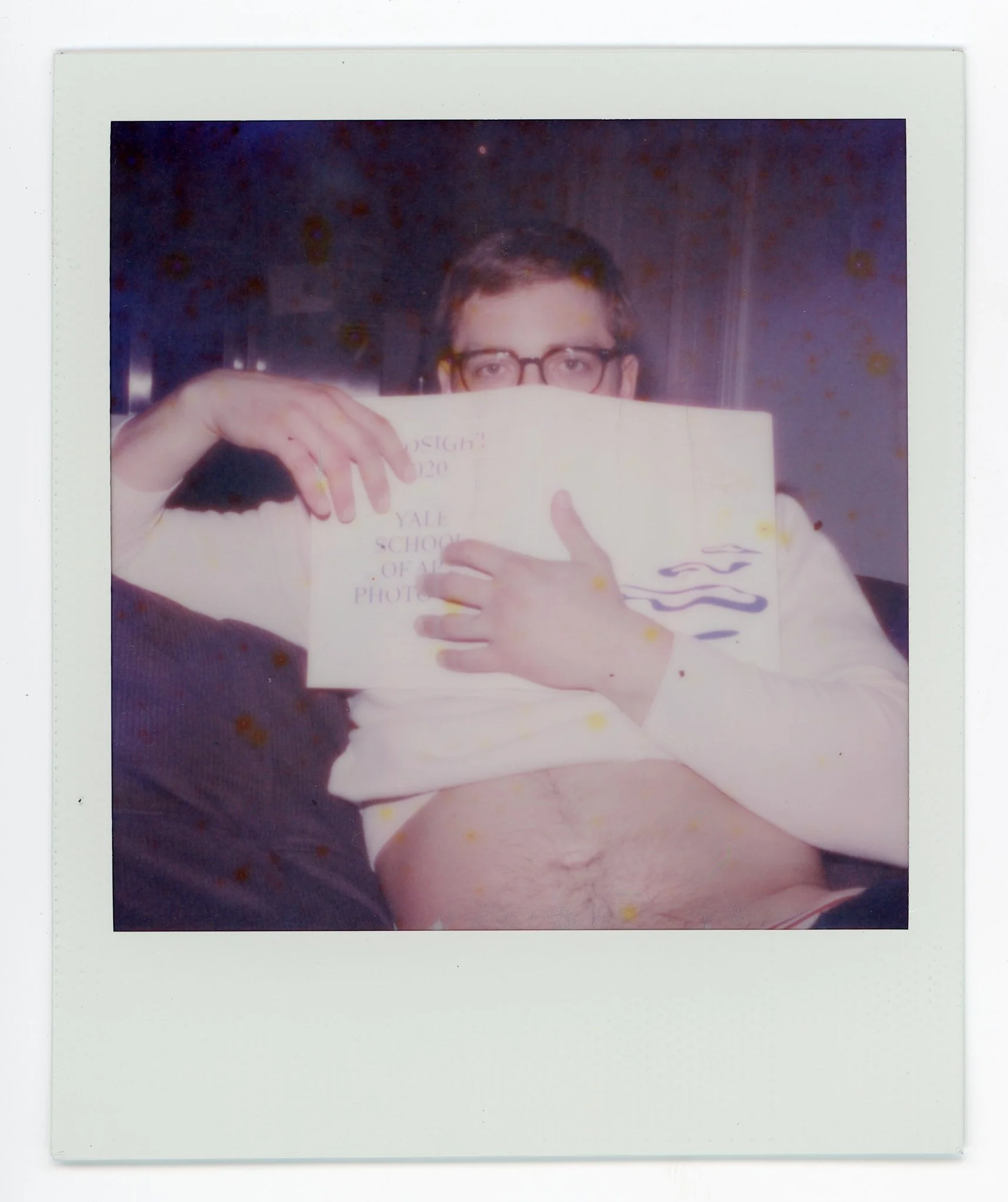 A young man wearing glasses holding a white paper or book in front of his face, partially obscuring it, with only his eyes visible. The person appears to be shirtless.