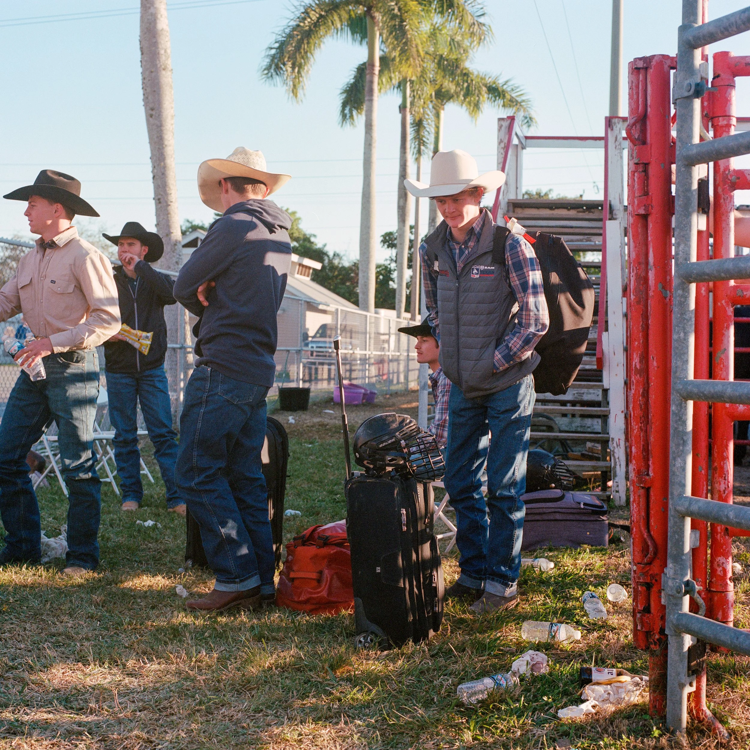 Group of young men wearing cowboy hats and casual clothes standing near a red metal gate at an outdoor event with palm trees in the background.
