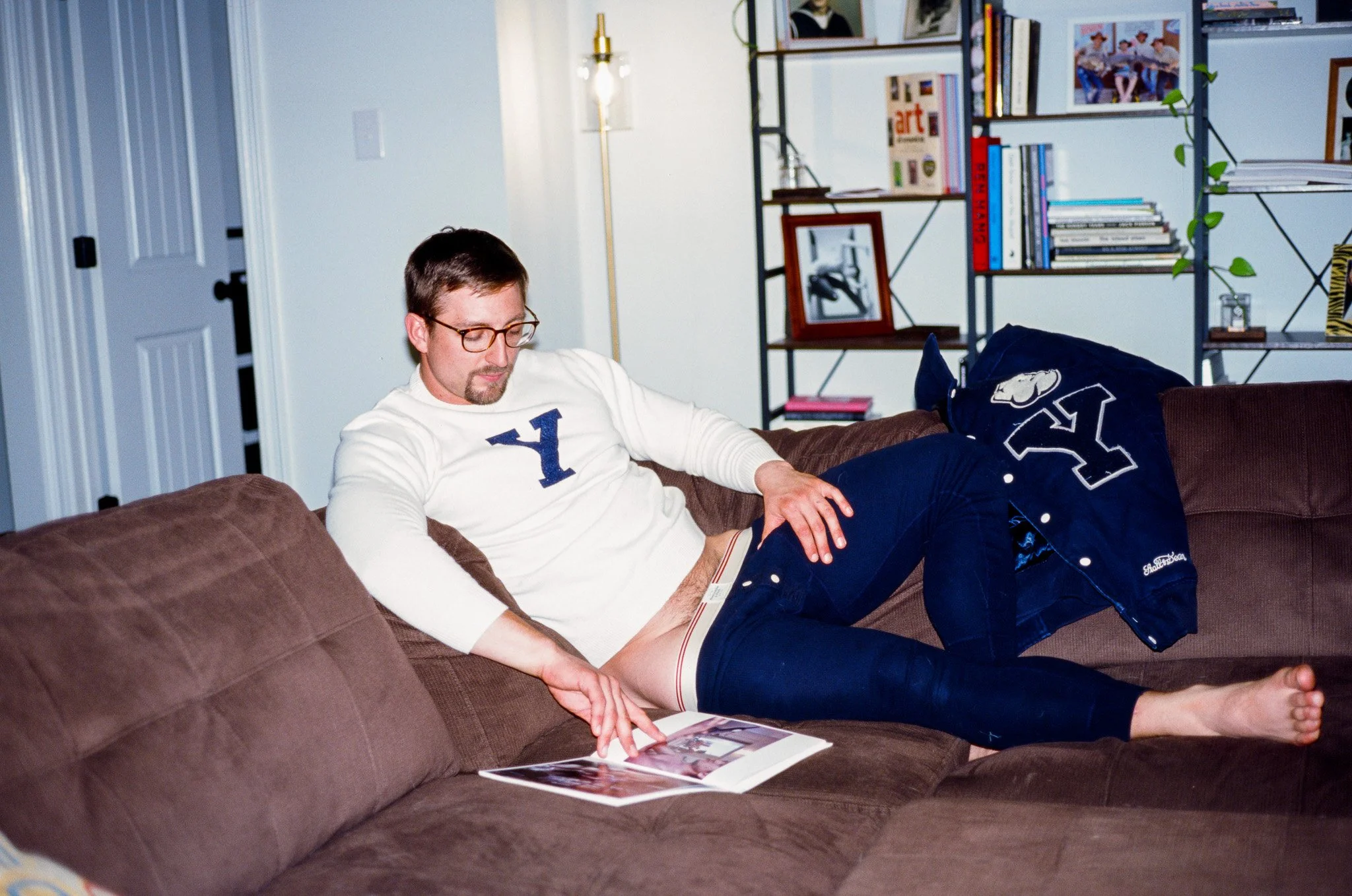 A man with glasses, a beard, and wearing a white long-sleeve shirt with a blue letter Y is lounging on a brown couch. He is looking down at a magazine. A blue letterman jacket with a large letter Y on the back is draped over the couch. Behind him is 