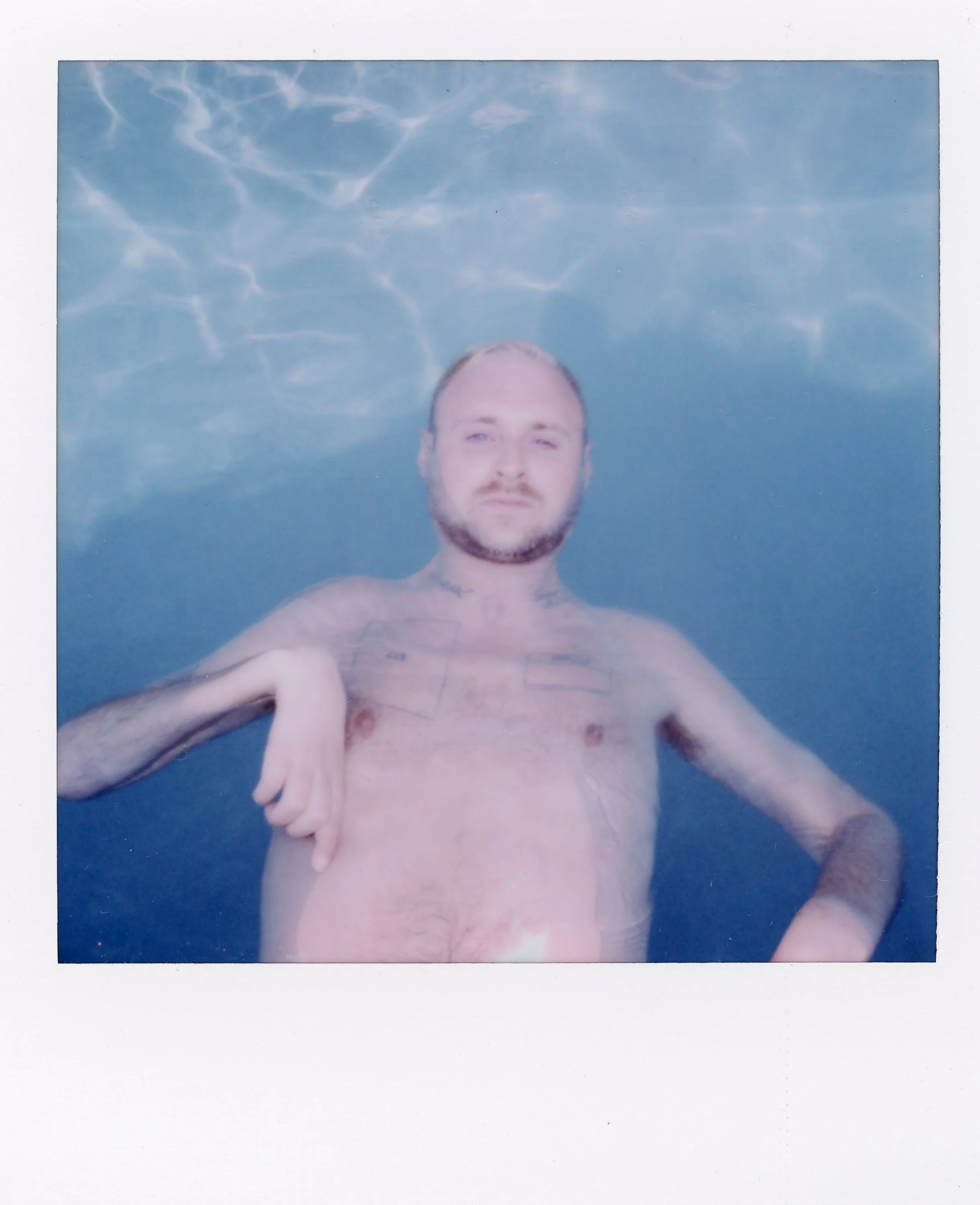 A man with a beard and tattoos in a swimming pool, with his arm resting on his chest and a calm expression, water and sky reflections visible above.