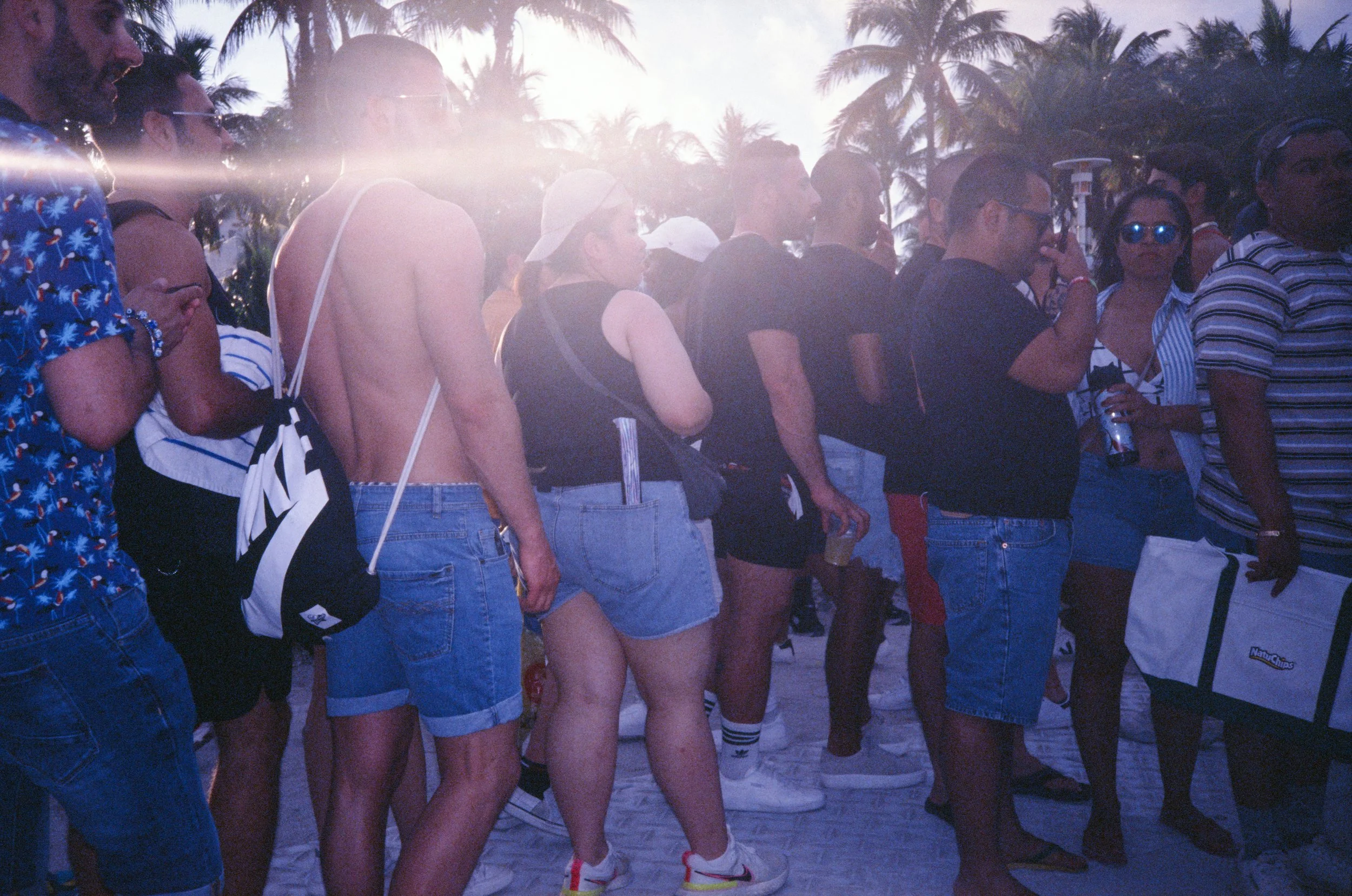 A line of people waiting outdoors with palm trees in the background, some wearing summer clothes, sunglasses, and headphones, during sunset.