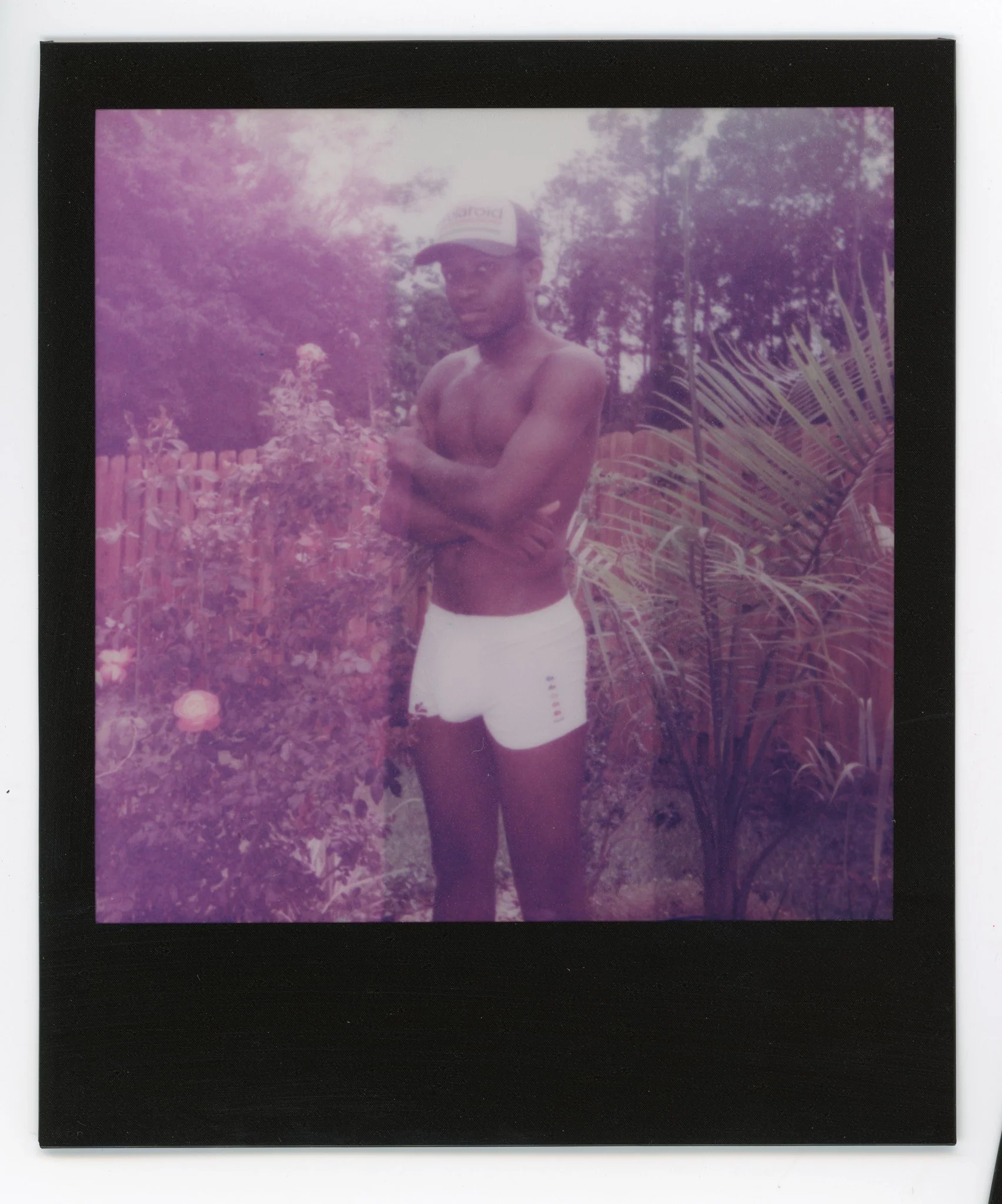 A shirtless young man wearing a cap and white shorts standing outdoors with arms crossed, surrounded by trees and plants.