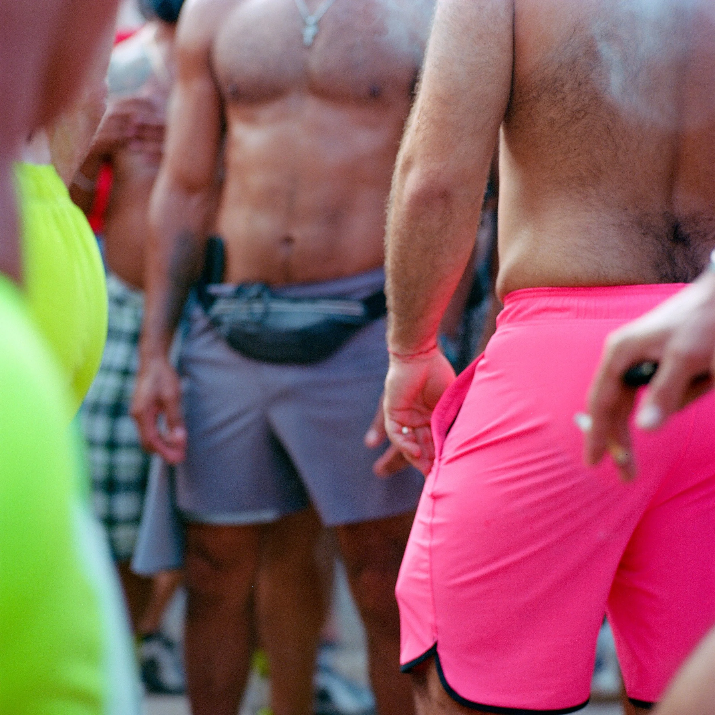Close-up of shirtless men in bright swim trunks, gathering outdoors.