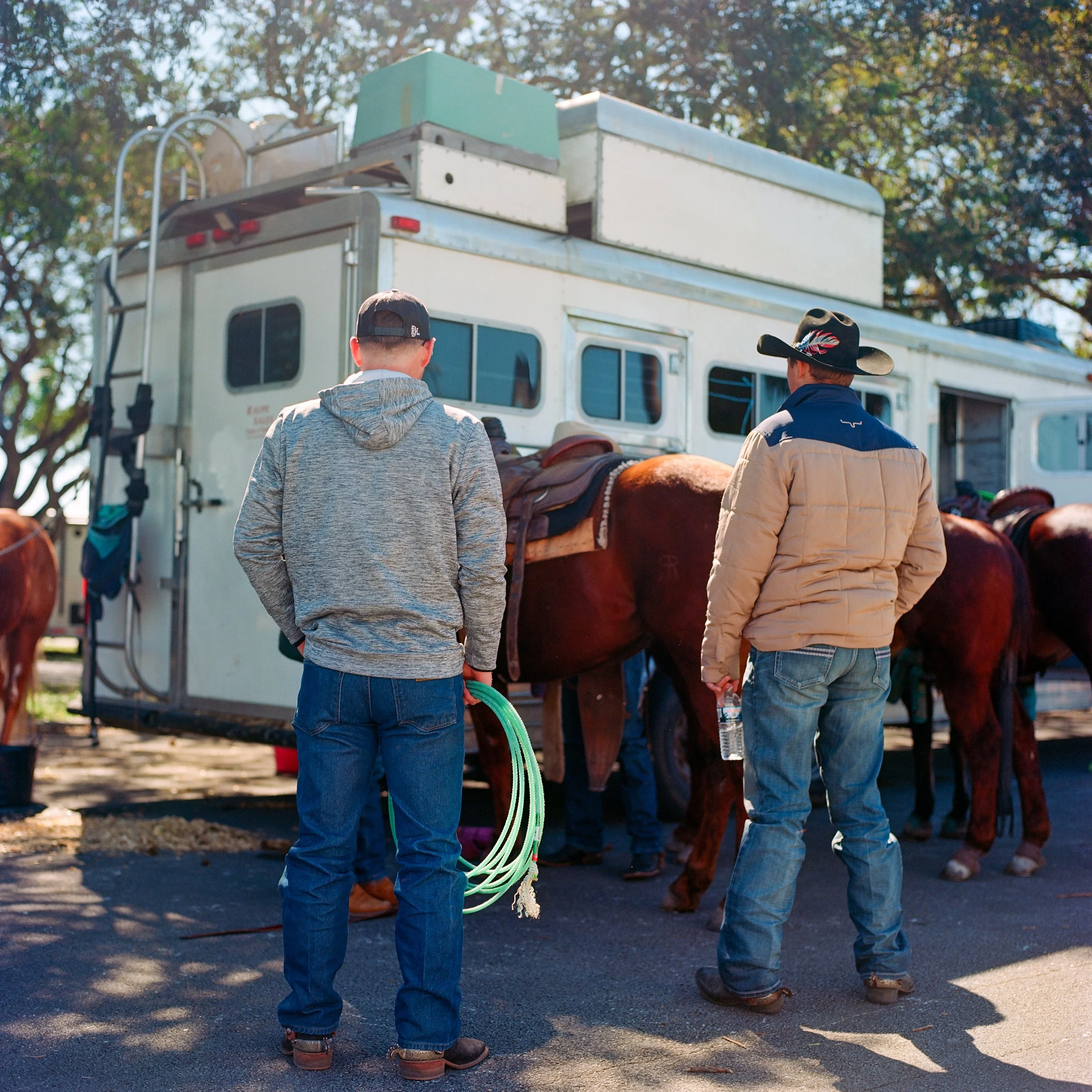 Two men in western attire standing near a horse trailer with horses; one holding a green hose, outdoors under trees.