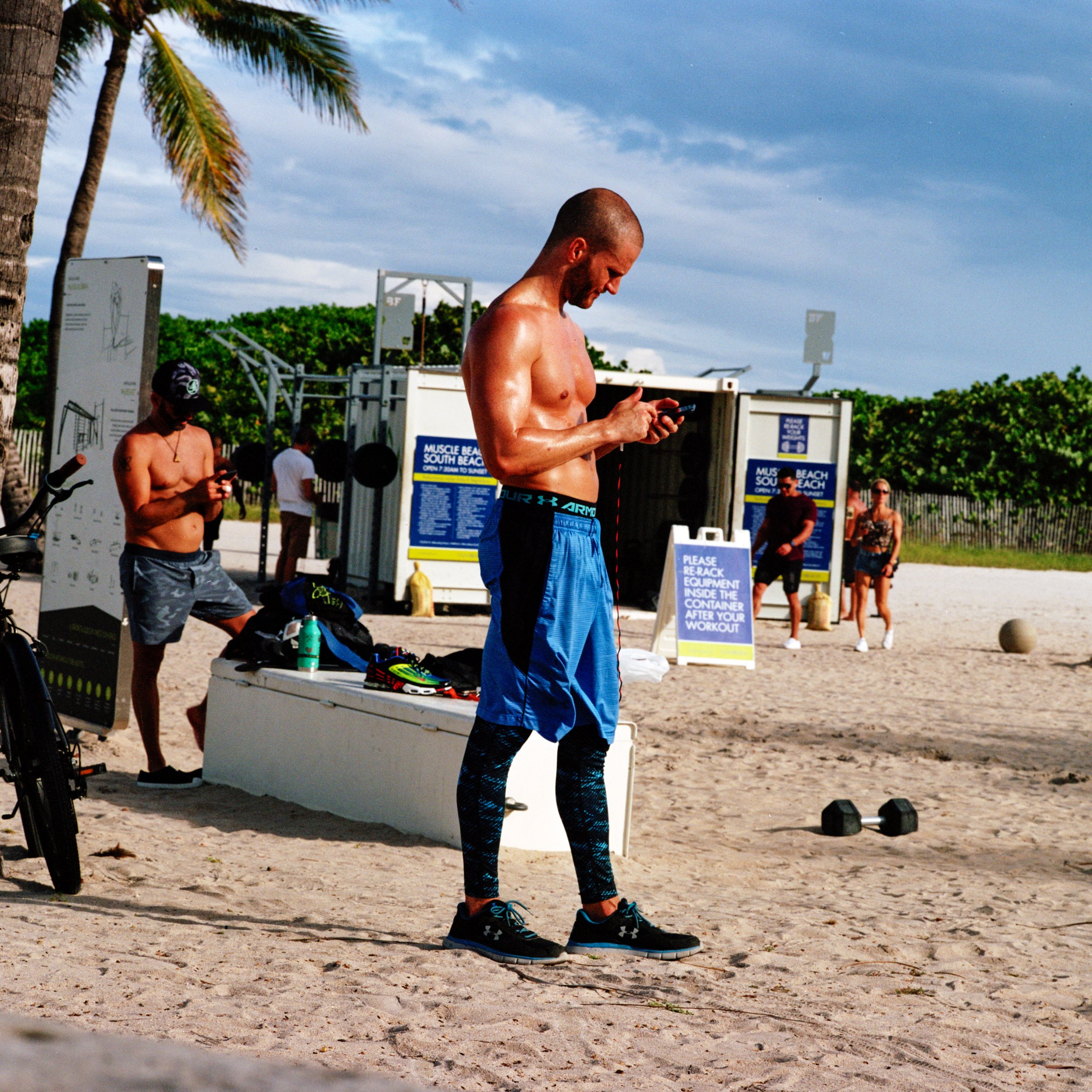 A shirtless man in athletic shorts and black running shoes standing on a sandy beach, looking at his phone. In the background, other people are also shirtless and casual, with tropical palm trees and gym equipment around, suggesting a beach workout o