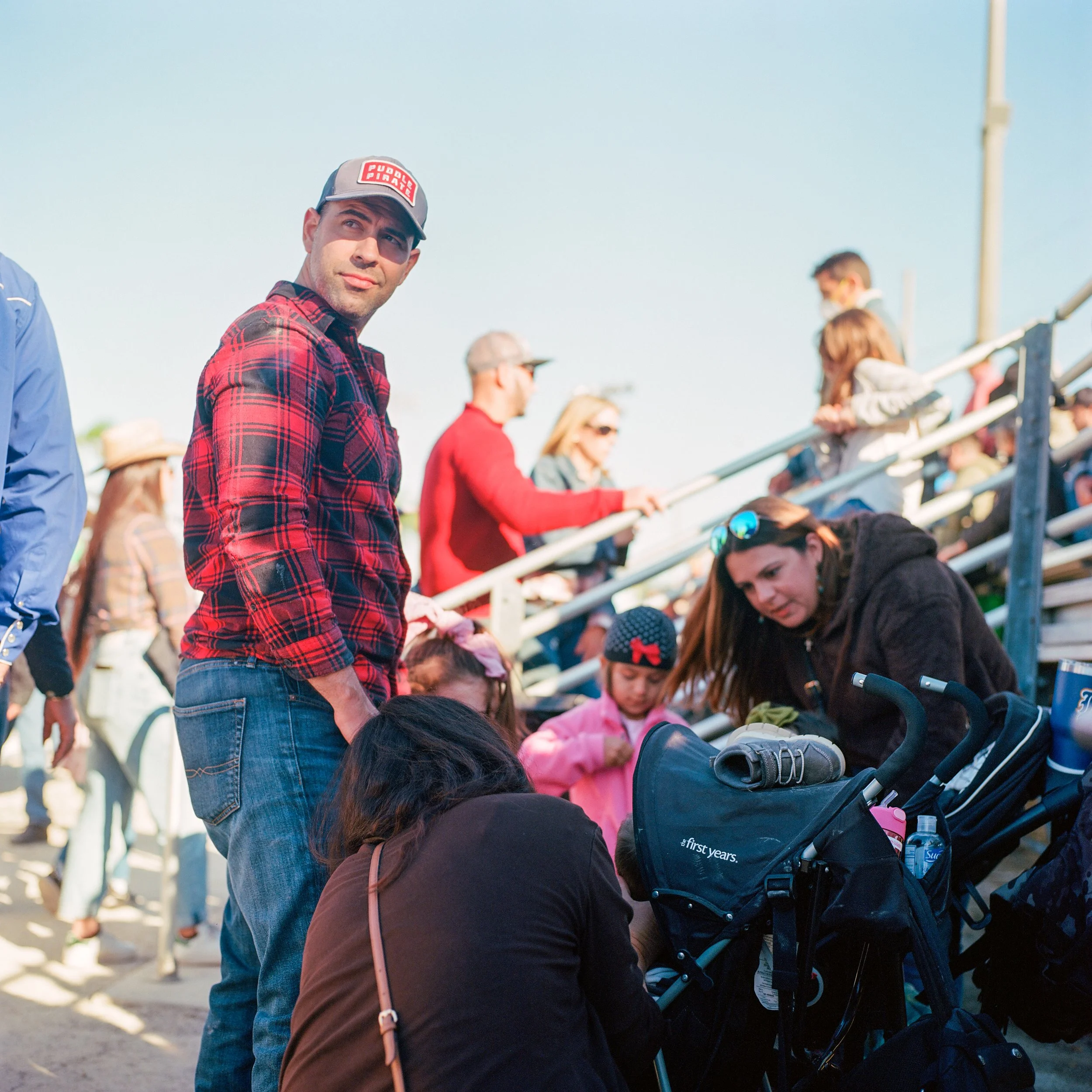 A man in a red plaid shirt and baseball cap standing in a crowd at an outdoor event, with people sitting on bleachers in the background.