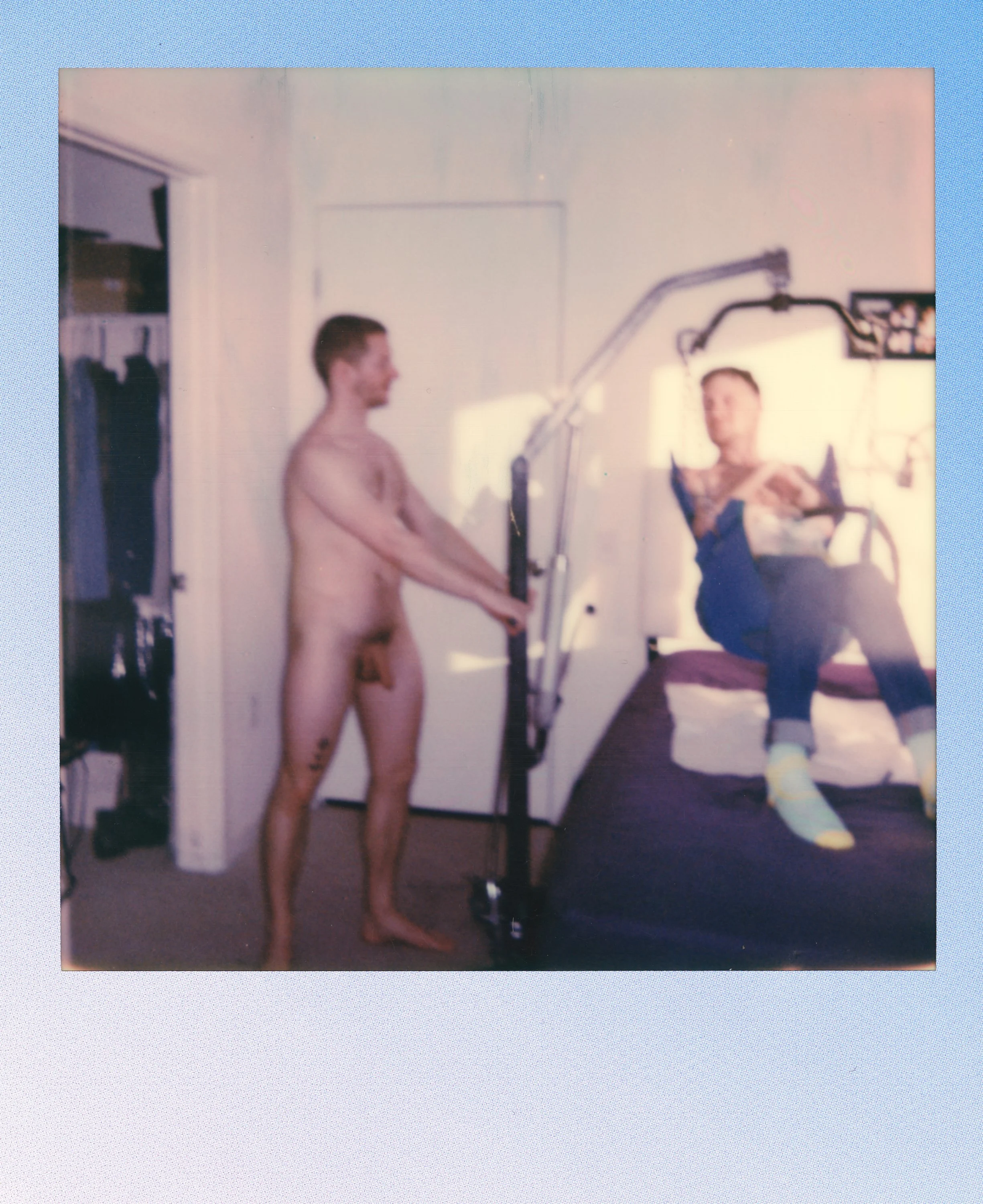 A person with tattoos on their leg and torso is lying in a hospital bed, while another person stands beside the bed, holding a medical device, in a room with a closet and a door.