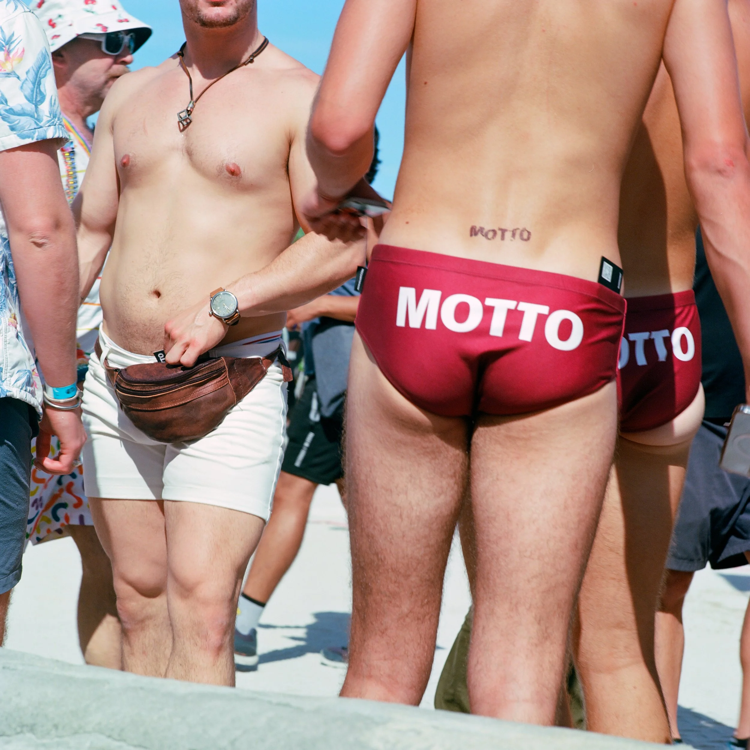 Several shirtless men wearing red underwear with the brand name 'MOTTO' printed on the back, standing on a sandy beach.